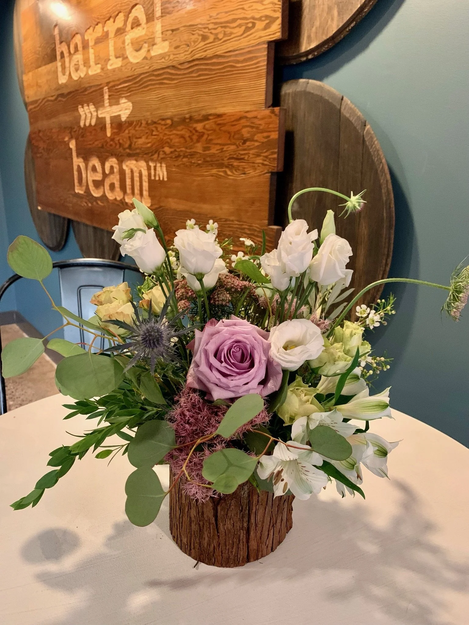 A wooden flower arrangement holder with a bouquet of white, pink, and purple flowers and green leaves, placed on a white surface with a wall and a decorated wooden sign in the background.