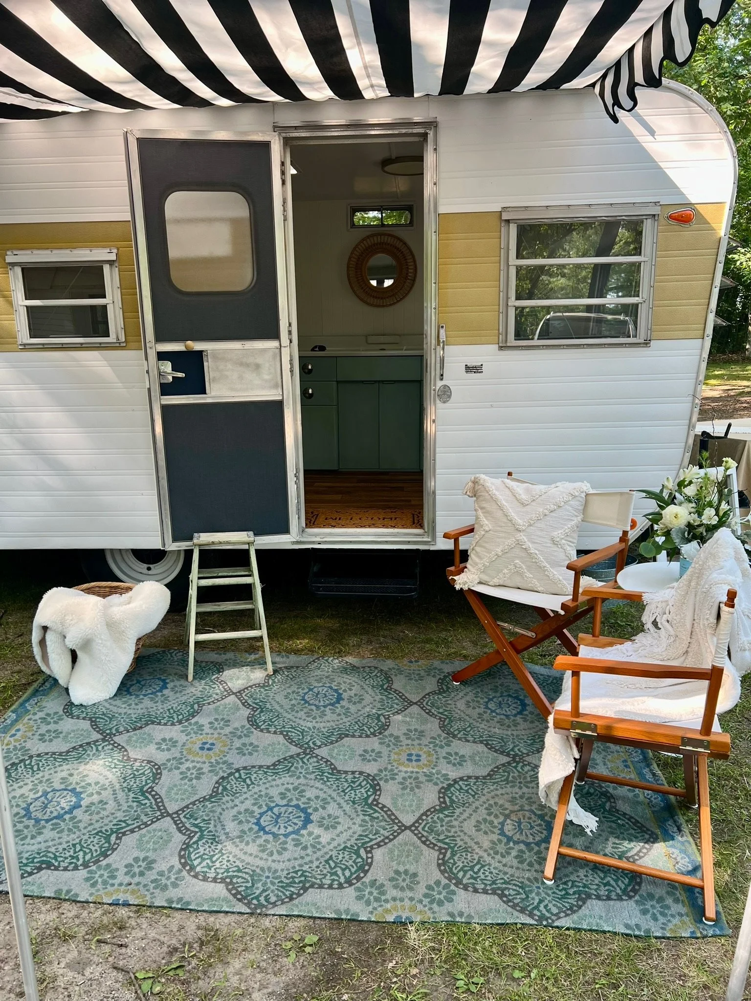 A vintage camper trailer set up outdoors with a striped awning, a patterned outdoor rug, a pair of wooden chairs with white cushions and throws, a potted plant, a towel, and a dog petting the rug. The camper has two small windows and an open door lea