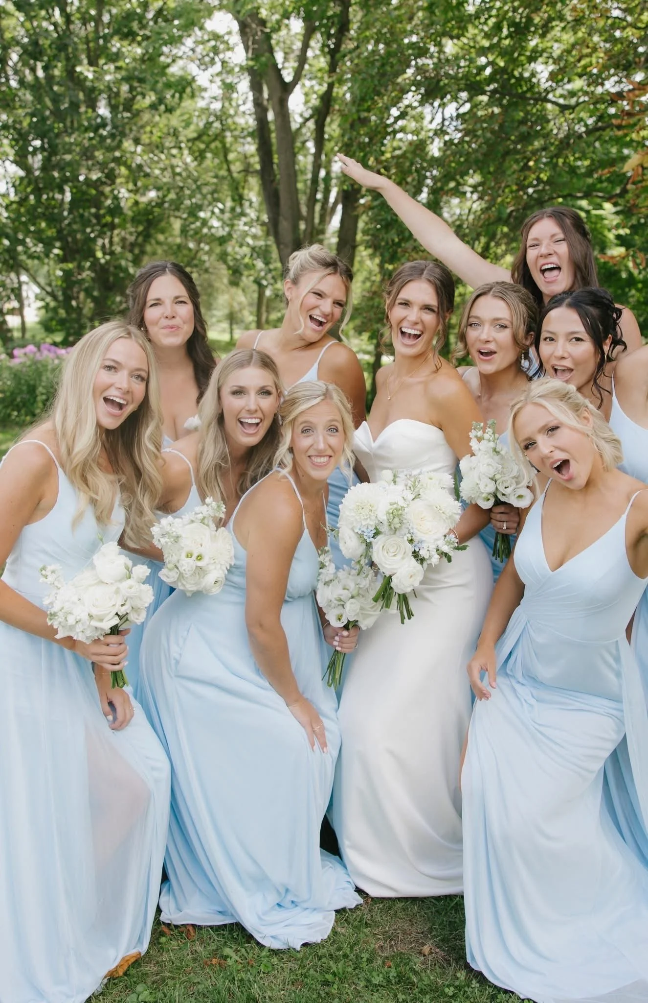 A group of nine women in matching white and light blue dresses posing outdoors on grass, holding white flower bouquets, smiling and laughing, with green trees in the background.