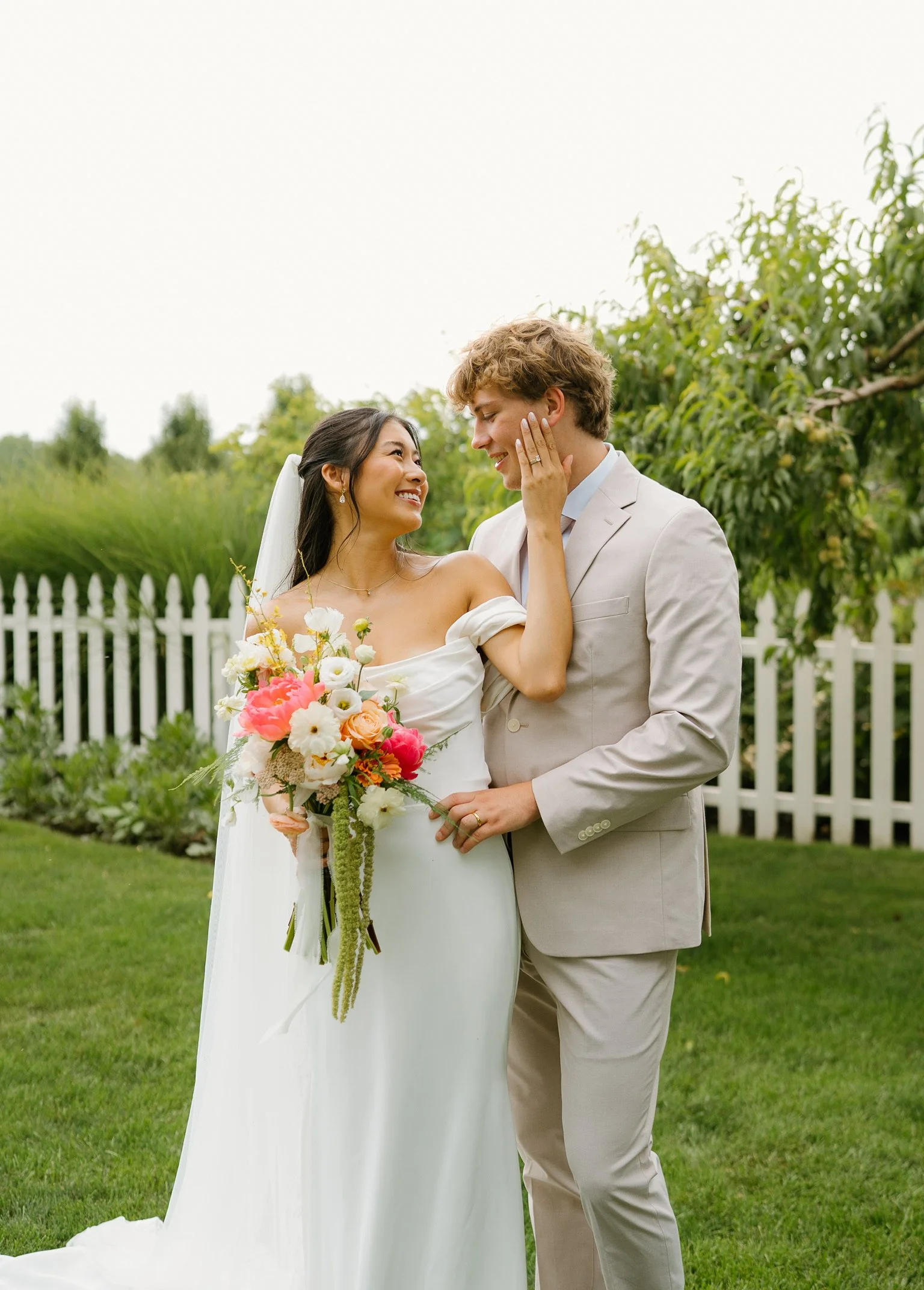 A bride and groom standing outdoors, smiling and looking at each other, with the bride touching the groom's face, surrounded by greenery and a white picket fence.
