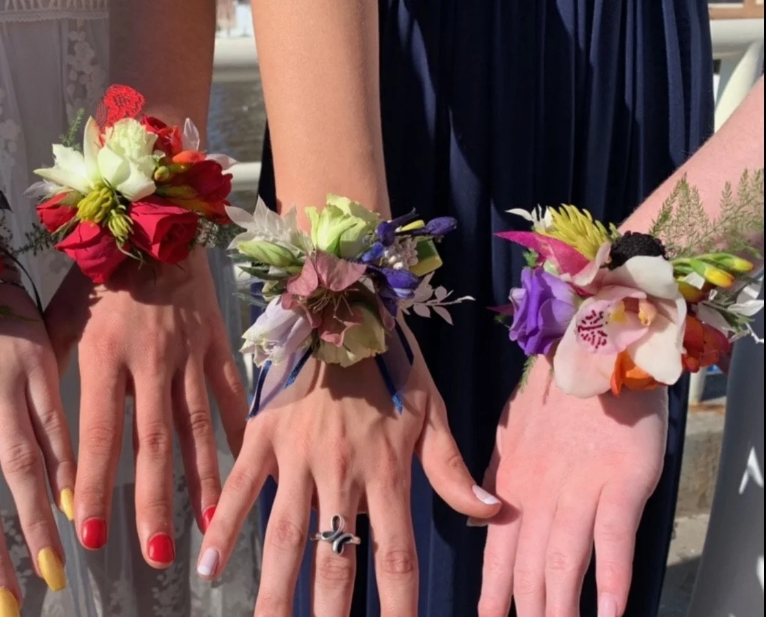 Three people wearing floral corsages on their wrists, showing their hands with red, white, and yellow painted nails.