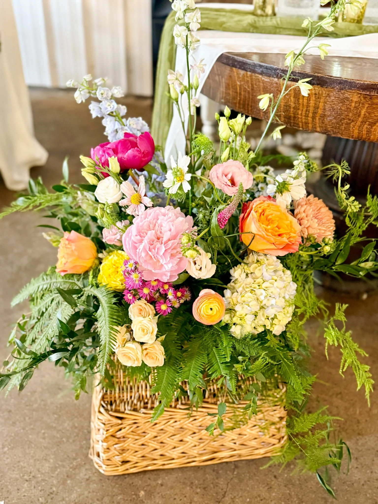 A large basket filled with a variety of colorful flowers, including pink, yellow, white, and orange blossoms, placed on a brown surface indoor.
