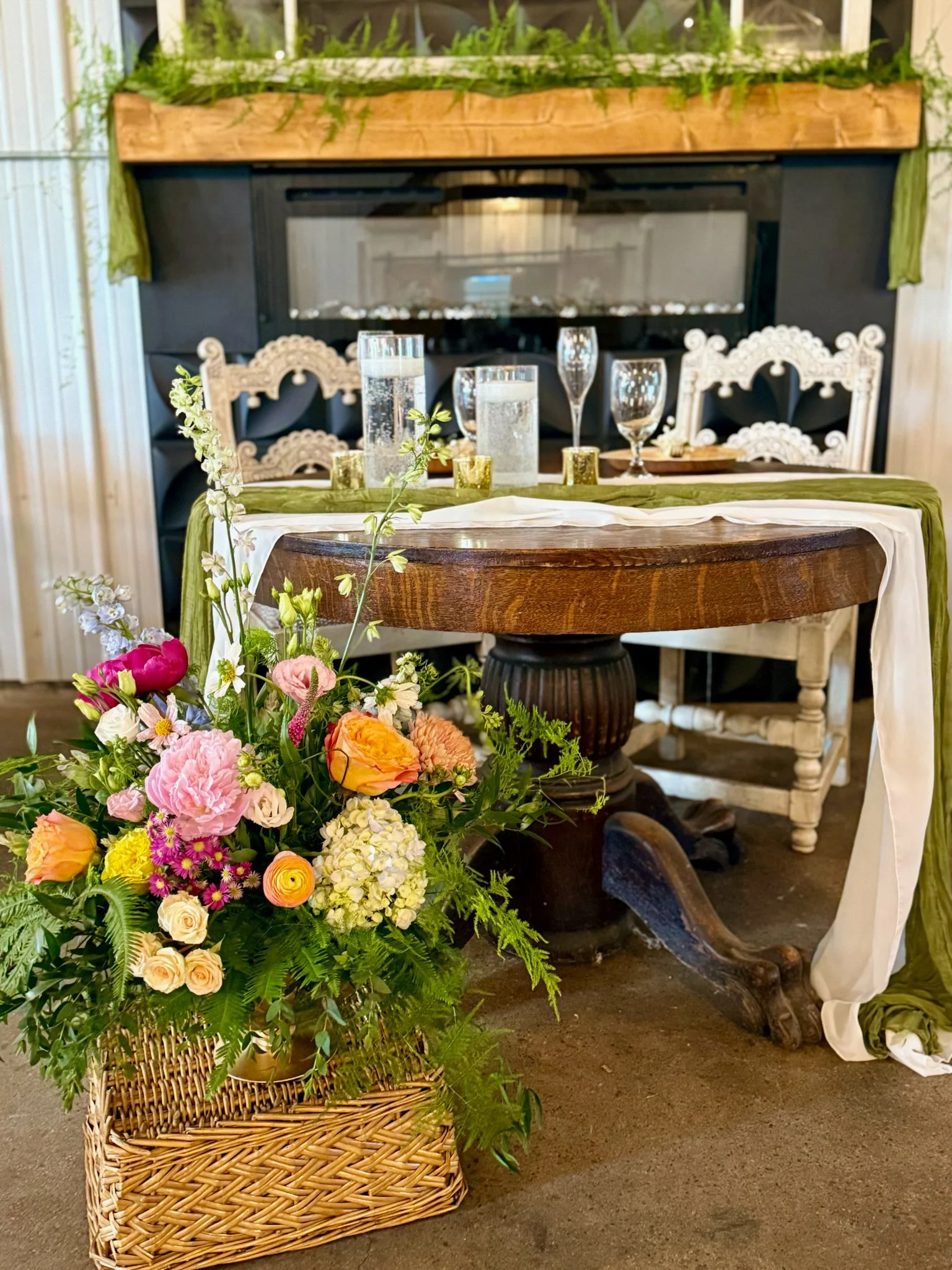A wicker basket filled with a colorful assortment of flowers, including pink, yellow, orange, and white blossoms, with green foliage. In the background, there is a decorated table with candles and glassware, and a floral arrangement on the table in f