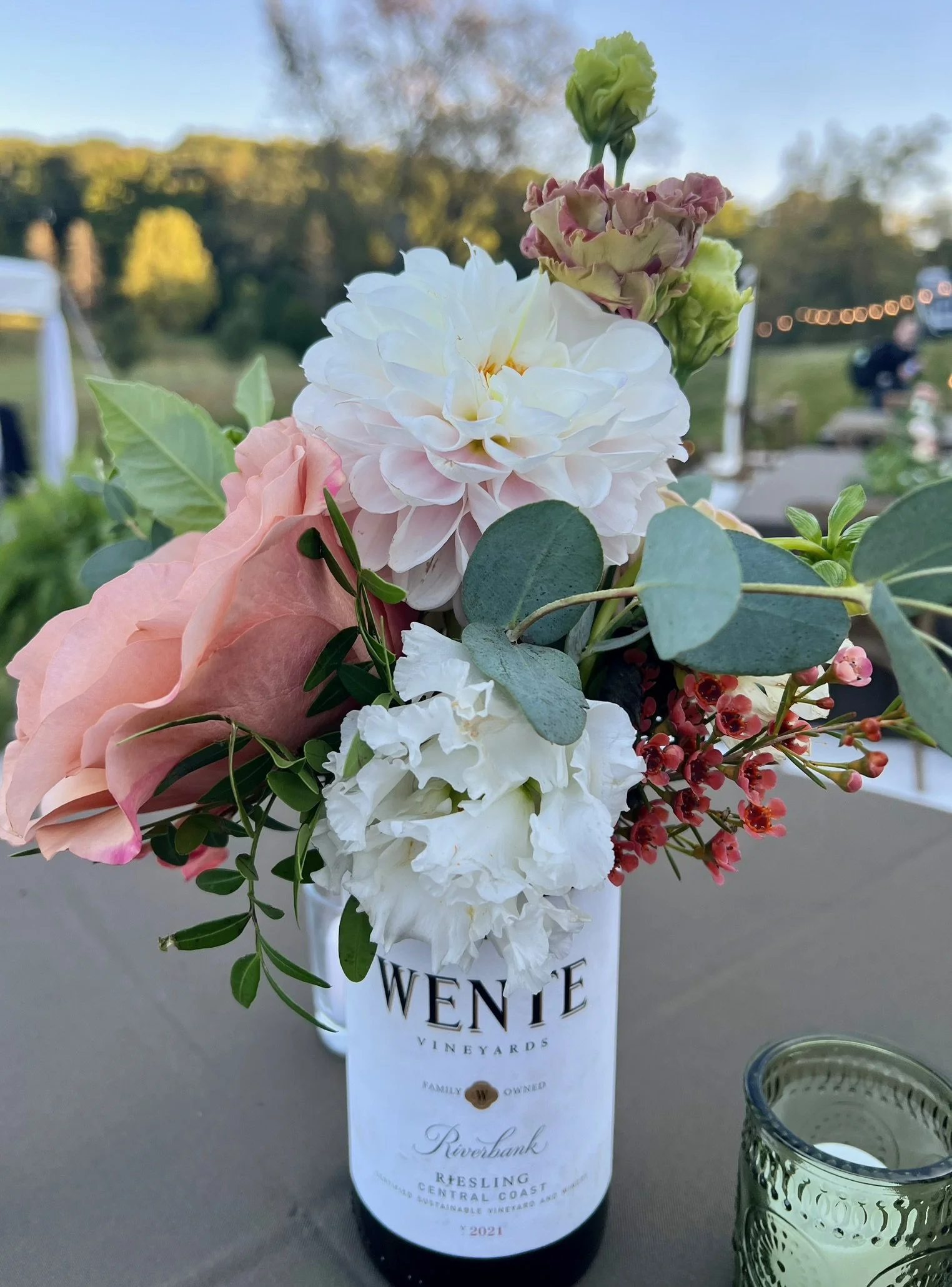 A wine bottle with floral decorations and a label reading 'Wente Vineyards Riverbank Riesling 2021' placed at an outdoor event. The background shows blurred trees and string lights.