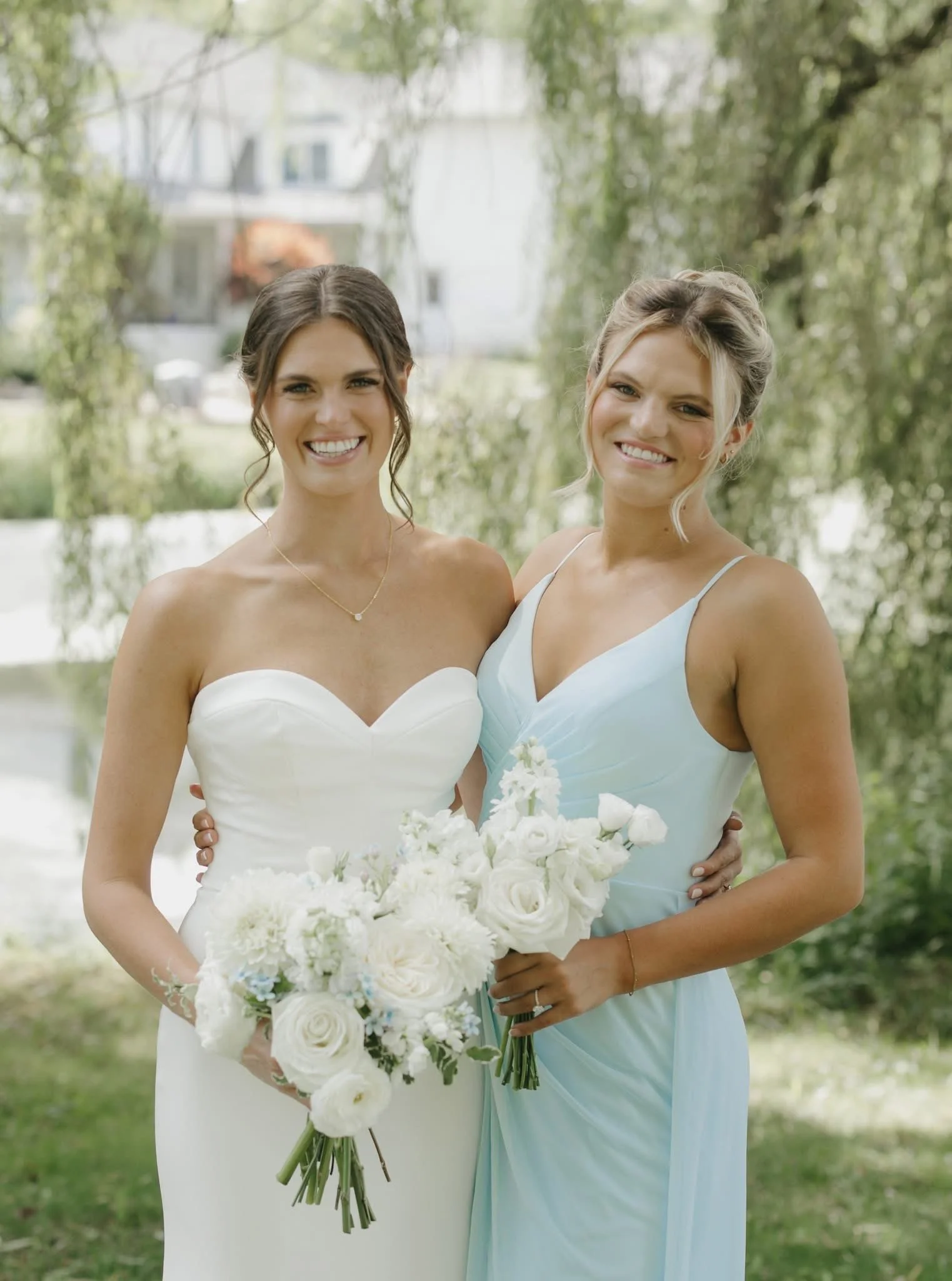 Two women in wedding dresses standing outdoors under green trees, smiling, holding bouquets of white flowers.
