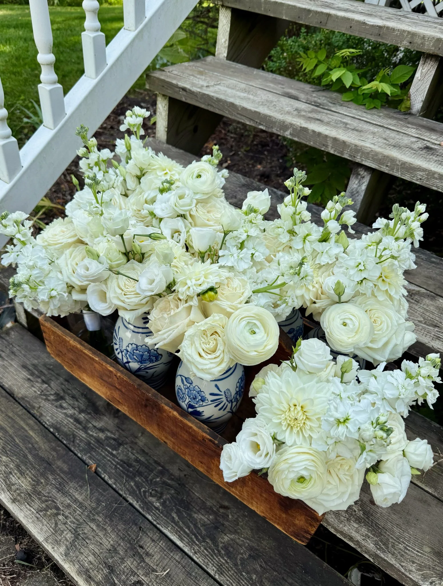 White flowers in decorative blue and white vases placed in a wooden container on a weathered wooden surface outdoors with stairs and greenery in the background.