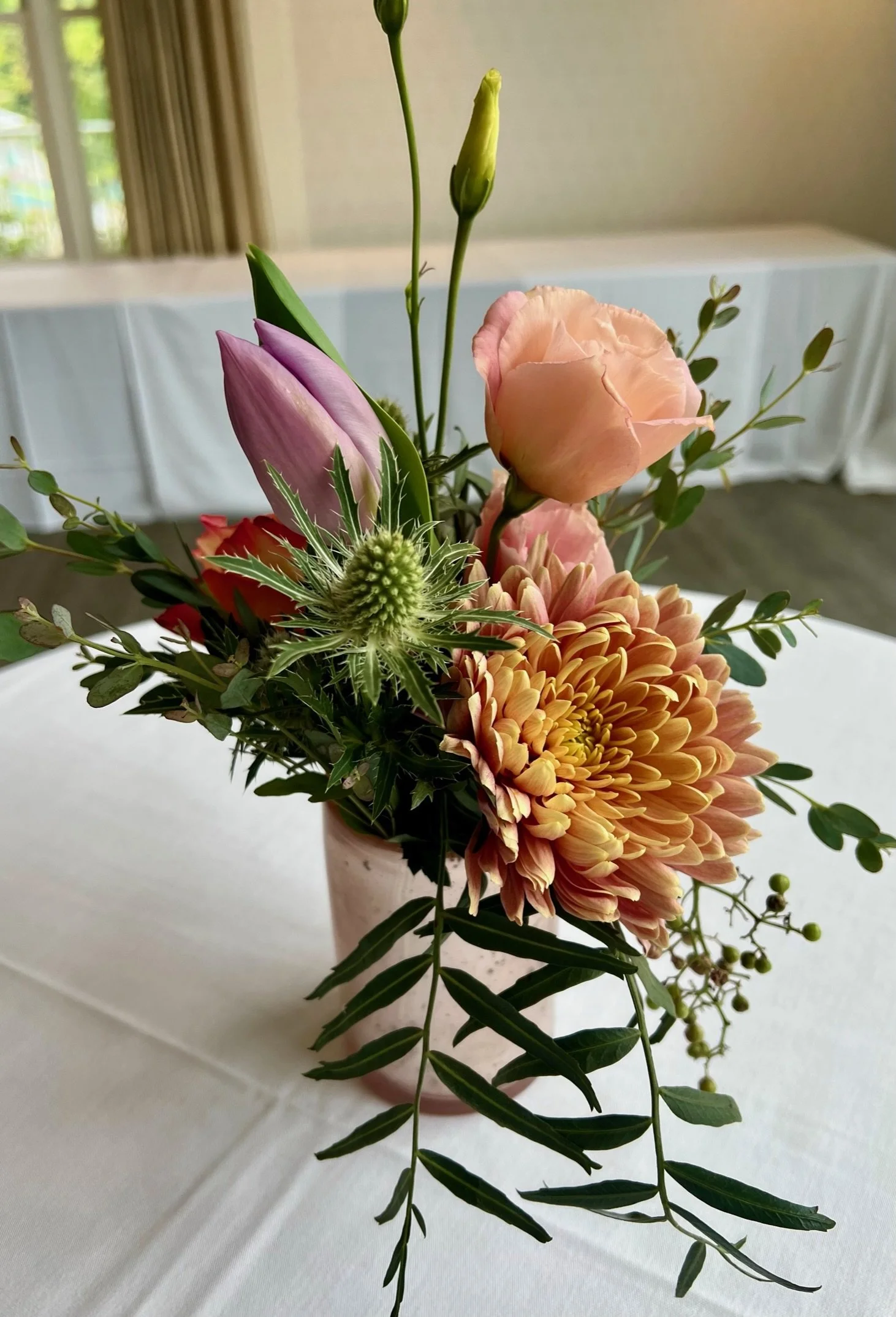 A bouquet of pink, peach, and purple flowers with greenery arranged in a vase on a white table, with a blurred room and window in the background.