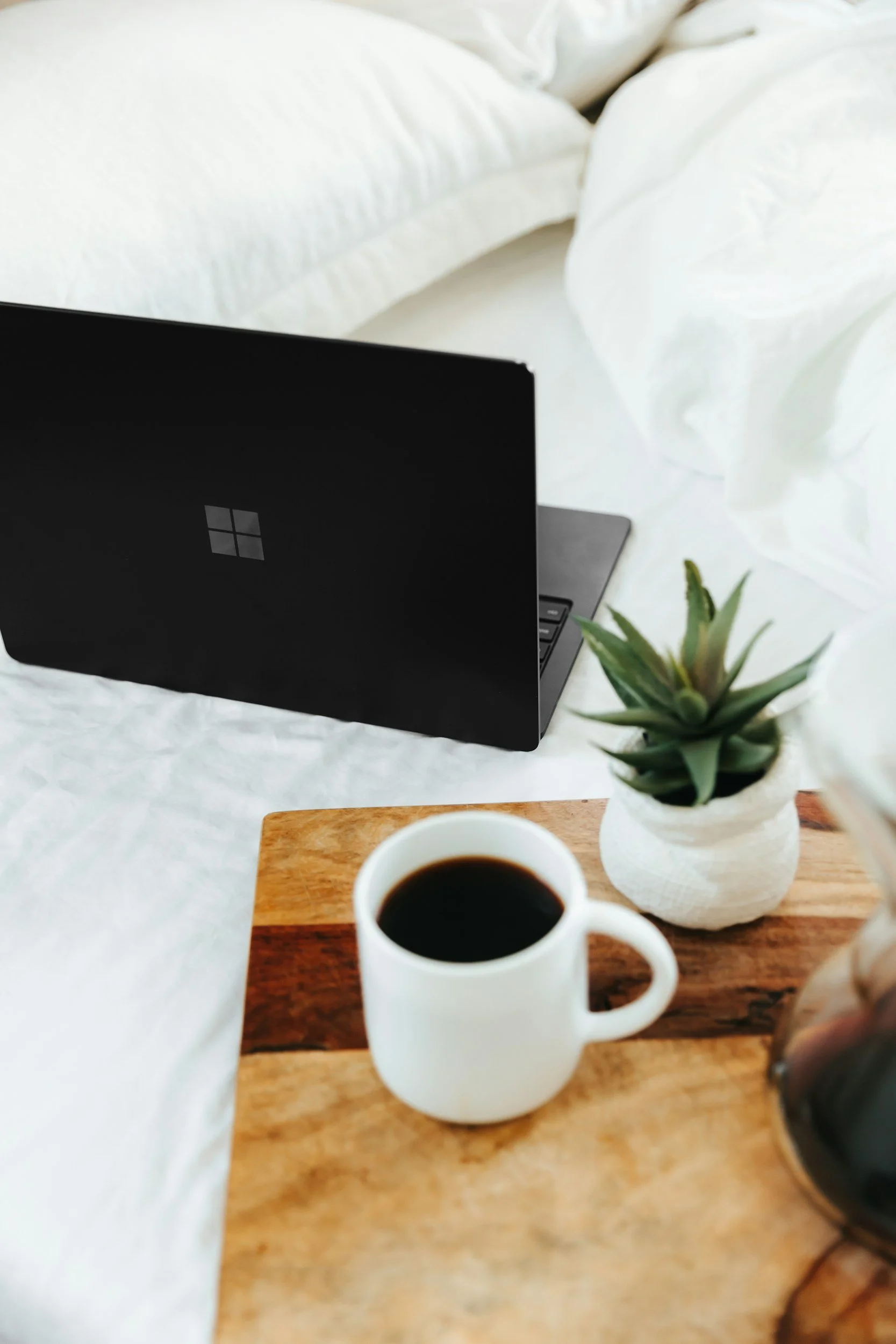 A picture of a laptop,plant, and cup of coffee beside a bed