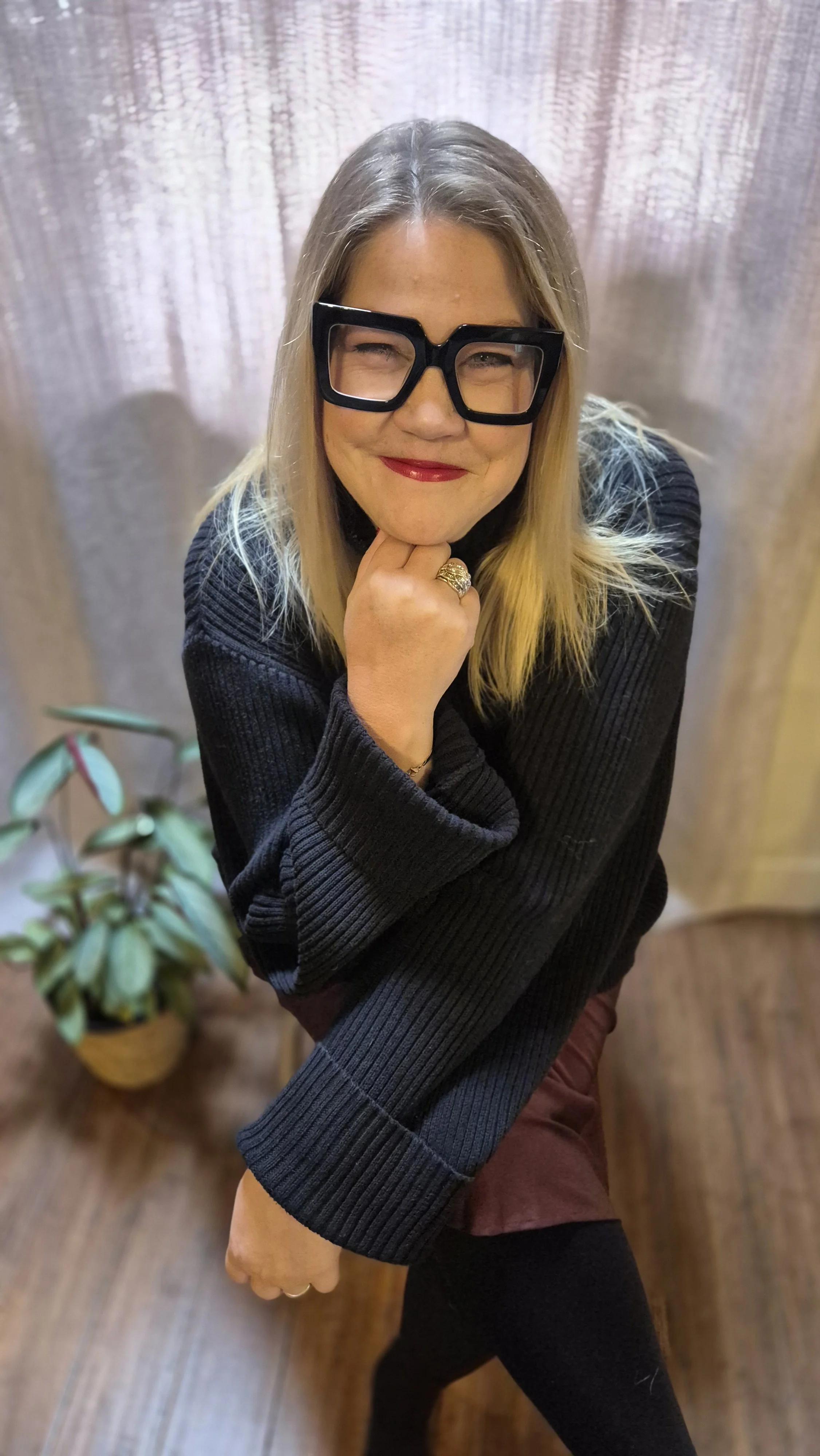 Susie Schwartz sitting on a stool in a black sweater and burgandy skirt. There is a plant beside her. She is wearing thick framed black glasses and is leaning on her hand and smiling