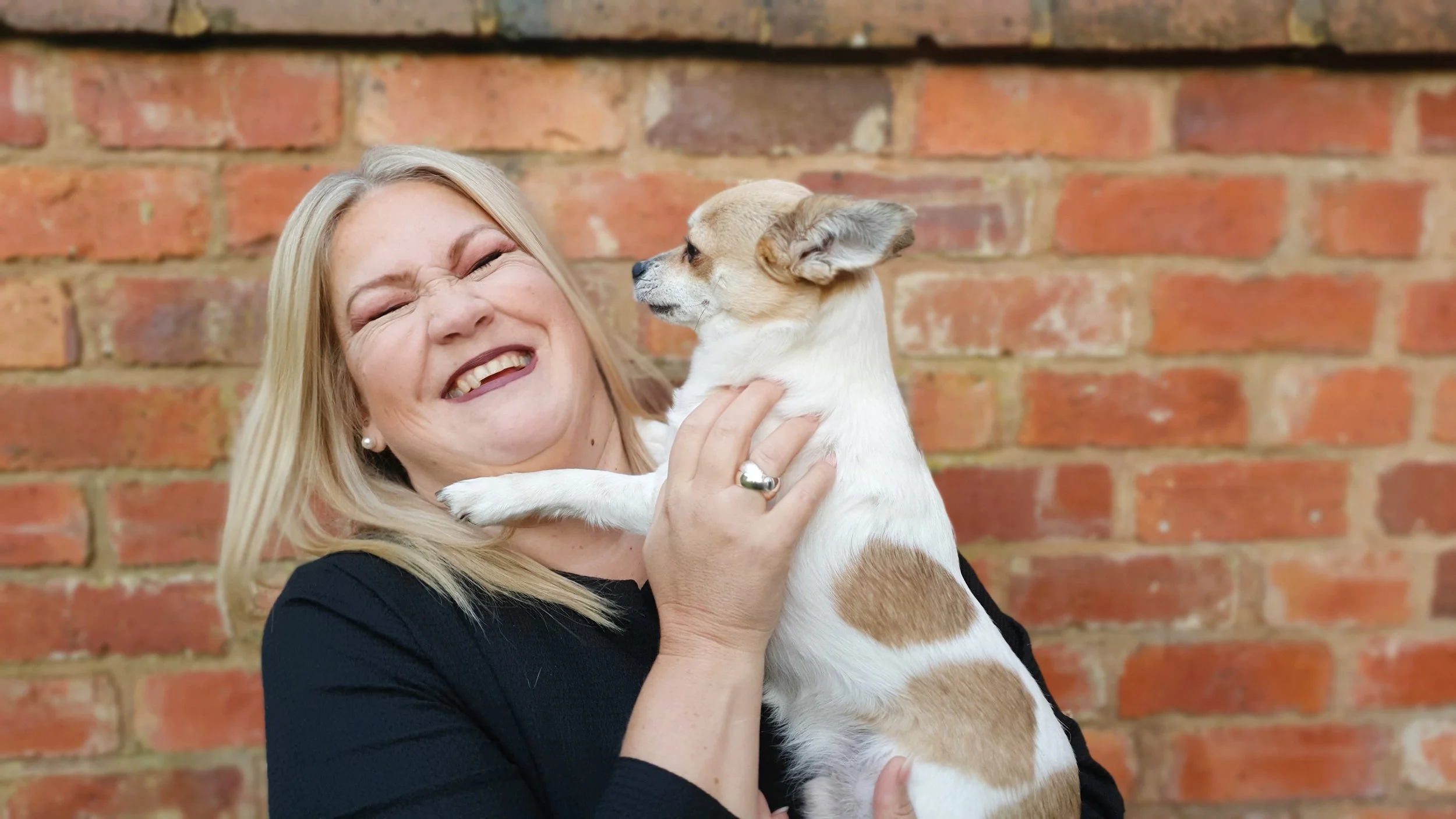 Susie Schwartz standing in front of a brick wall holding her chihuahua Carlos and laughing
