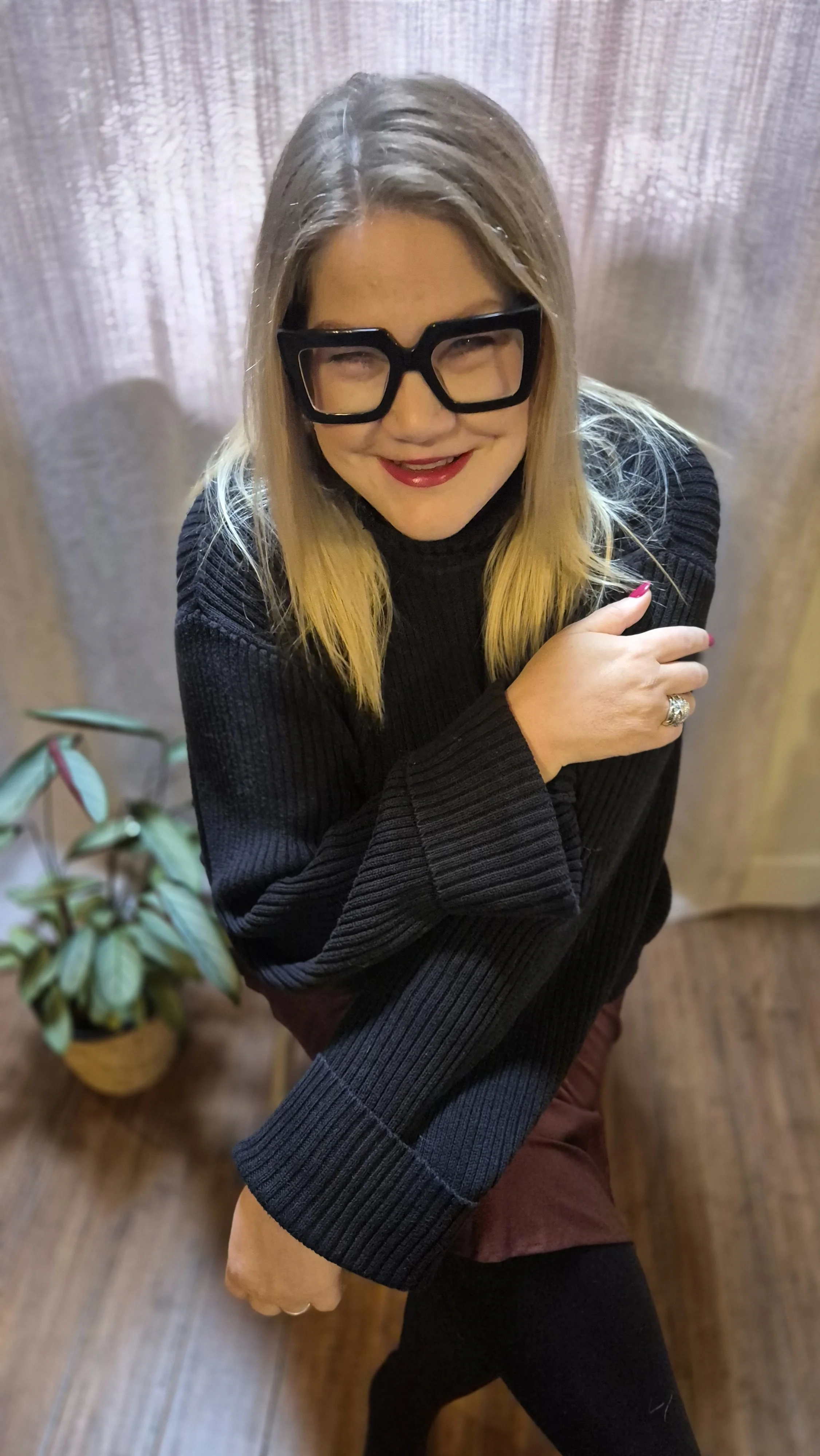 Susie Schwartz sitting on a stool beside a plant, wearing funky black glasses