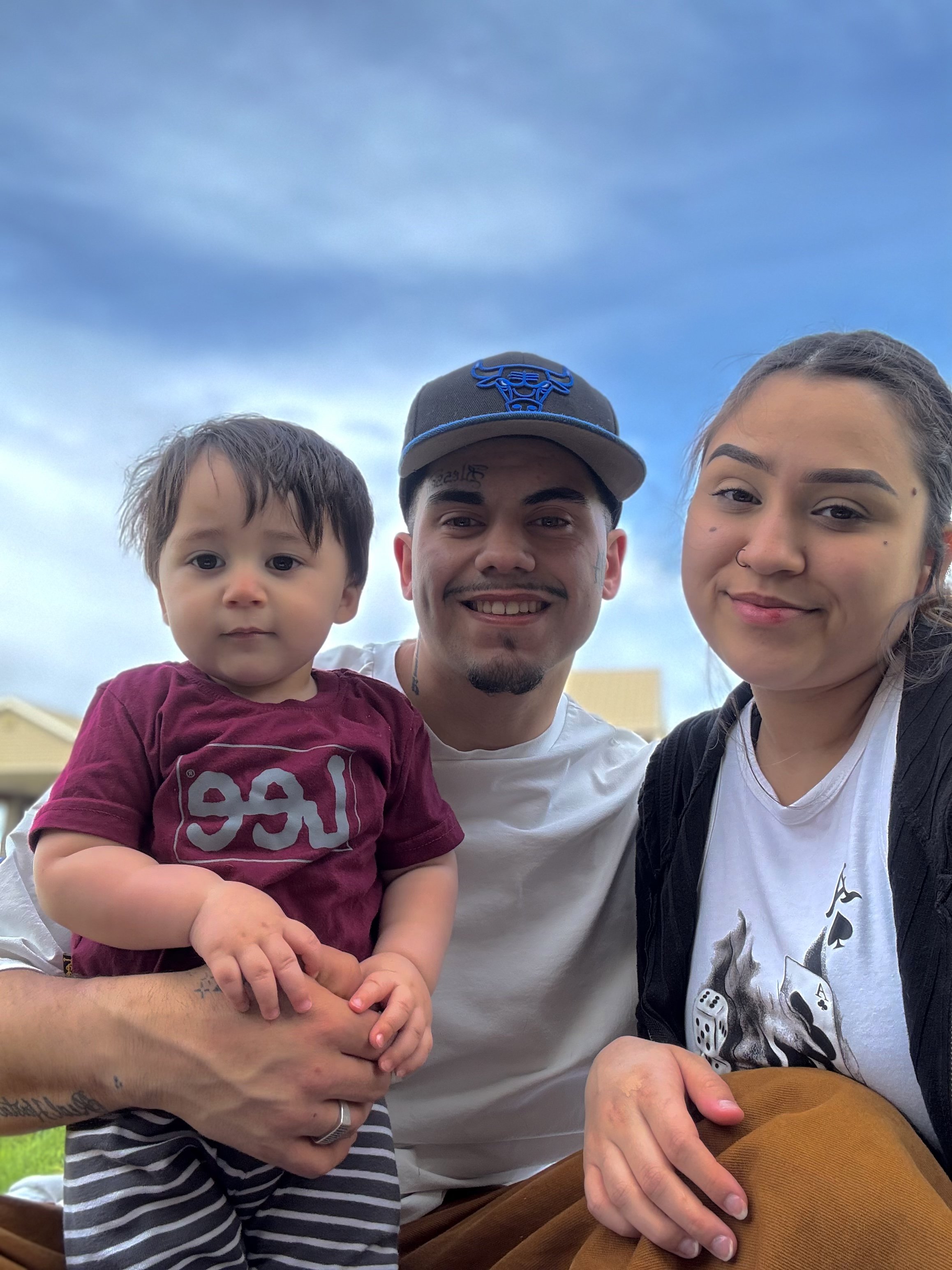 A man is holding a young child, and a woman is sitting next to him. They are outdoors under a cloudy sky.