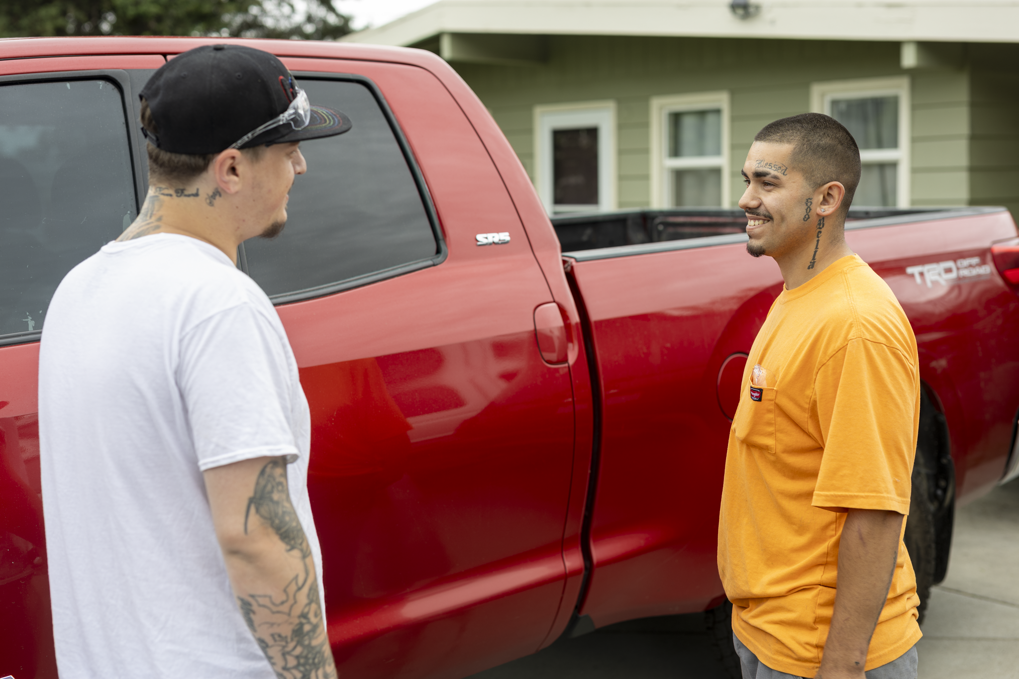 Two men facing each other and smiling, standing outdoors next to a red pickup truck, with a house in the background.