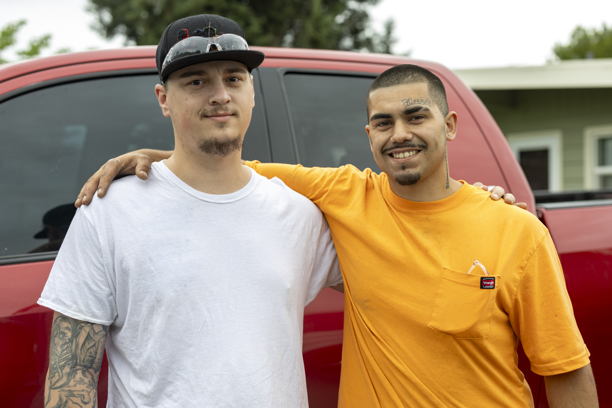 Two young men standing outside in front of a red vehicle, smiling and with arms around each other's shoulders. One is wearing a white t-shirt and a black cap with sunglasses, the other is wearing an orange shirt with tattoos on his face and neck.