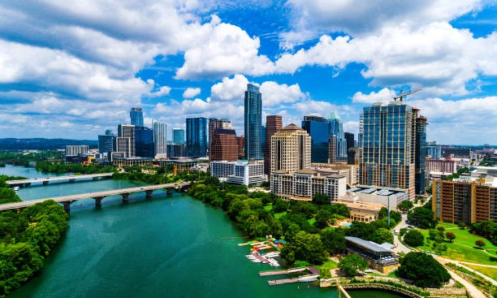 Aerial view of a city skyline with tall buildings, a river with bridges, and green parks under a partly cloudy sky.