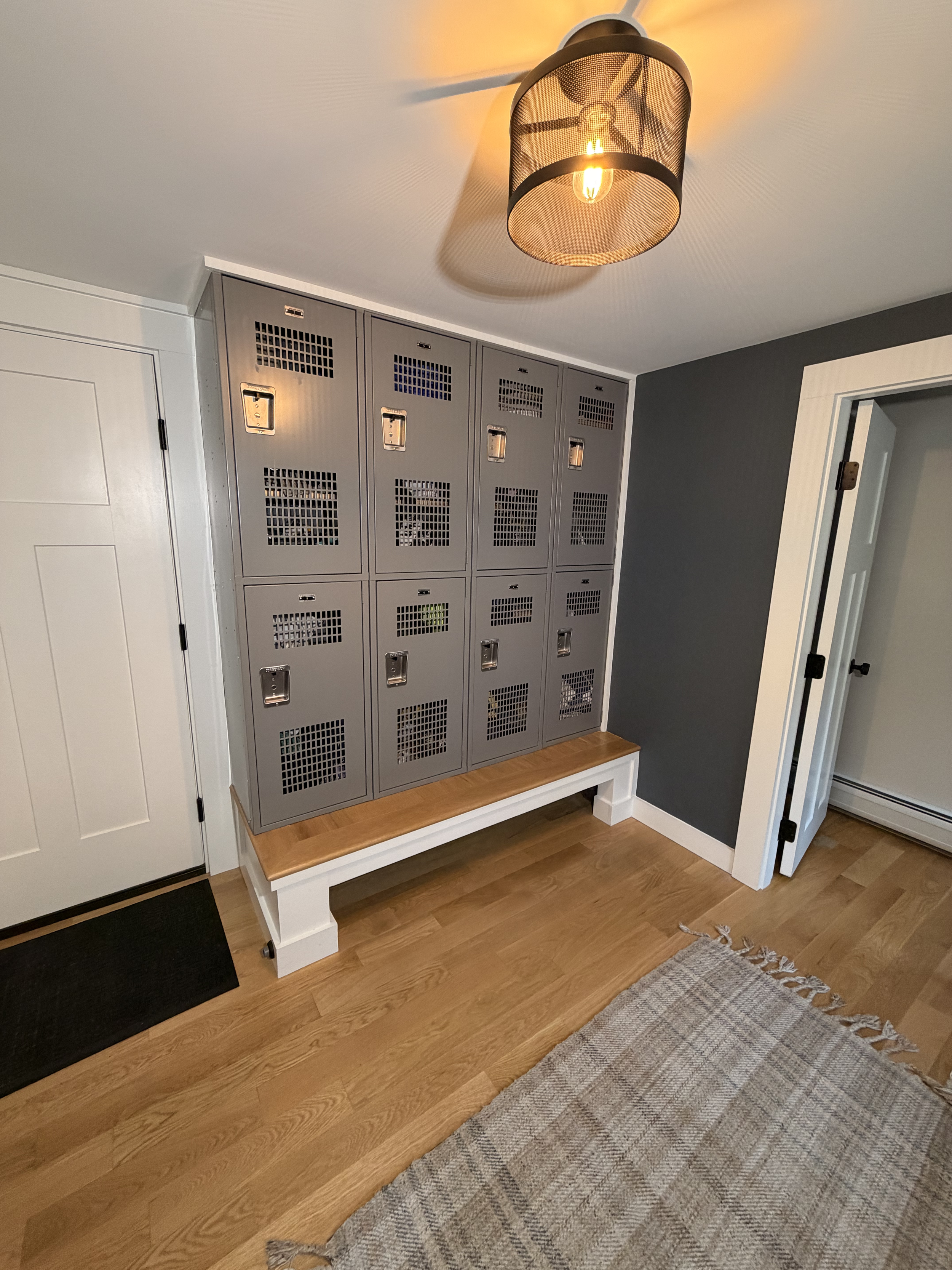 Gray metal lockers with ventilation holes, a handle on each door, and a rectangular lock. They are placed against a wall with a dark gray accent and sitting on a wooden bench with a white frame. Nearby is a green mat and a beige woven rug on a hardwood floor. The room has a ceiling mounted black cage light fixture with a visible filament bulb and white trim around the door frames.