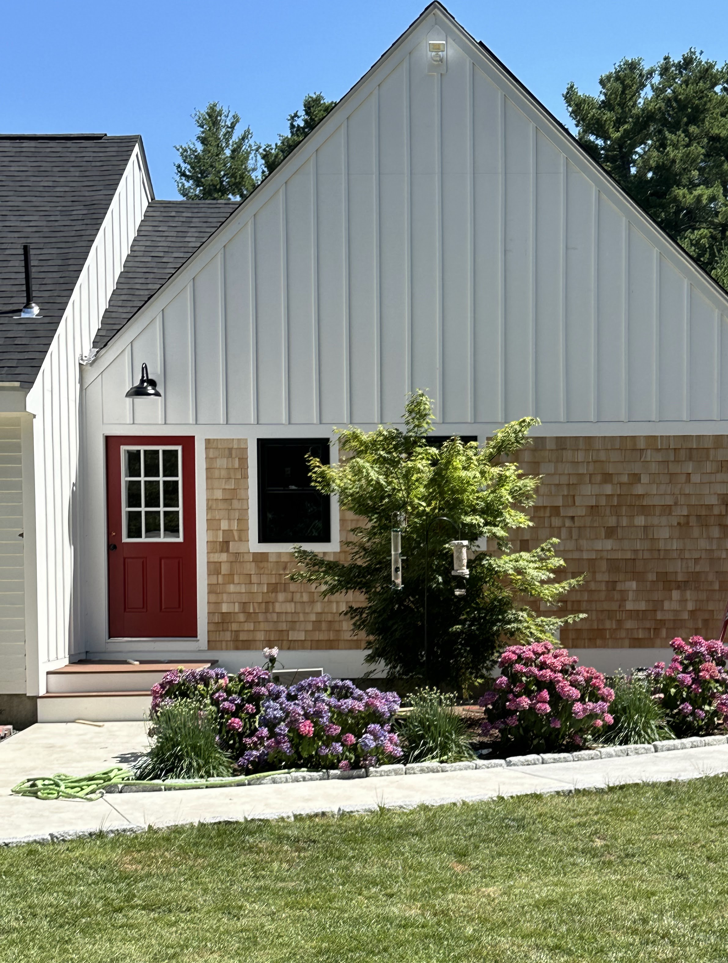 Front view of a house with a sloped roof, white siding, a red door, and a large window. There are colorful flowers and a small tree in the front yard, with a green lawn and a sidewalk.