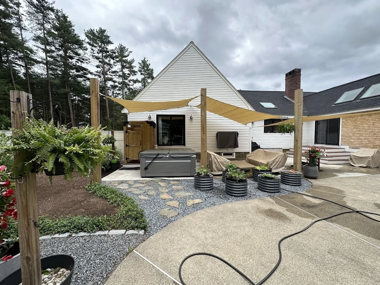 Backyard patio with a hot tub, covered furniture, and decorative planters, with a house and trees in the background under a cloudy sky.