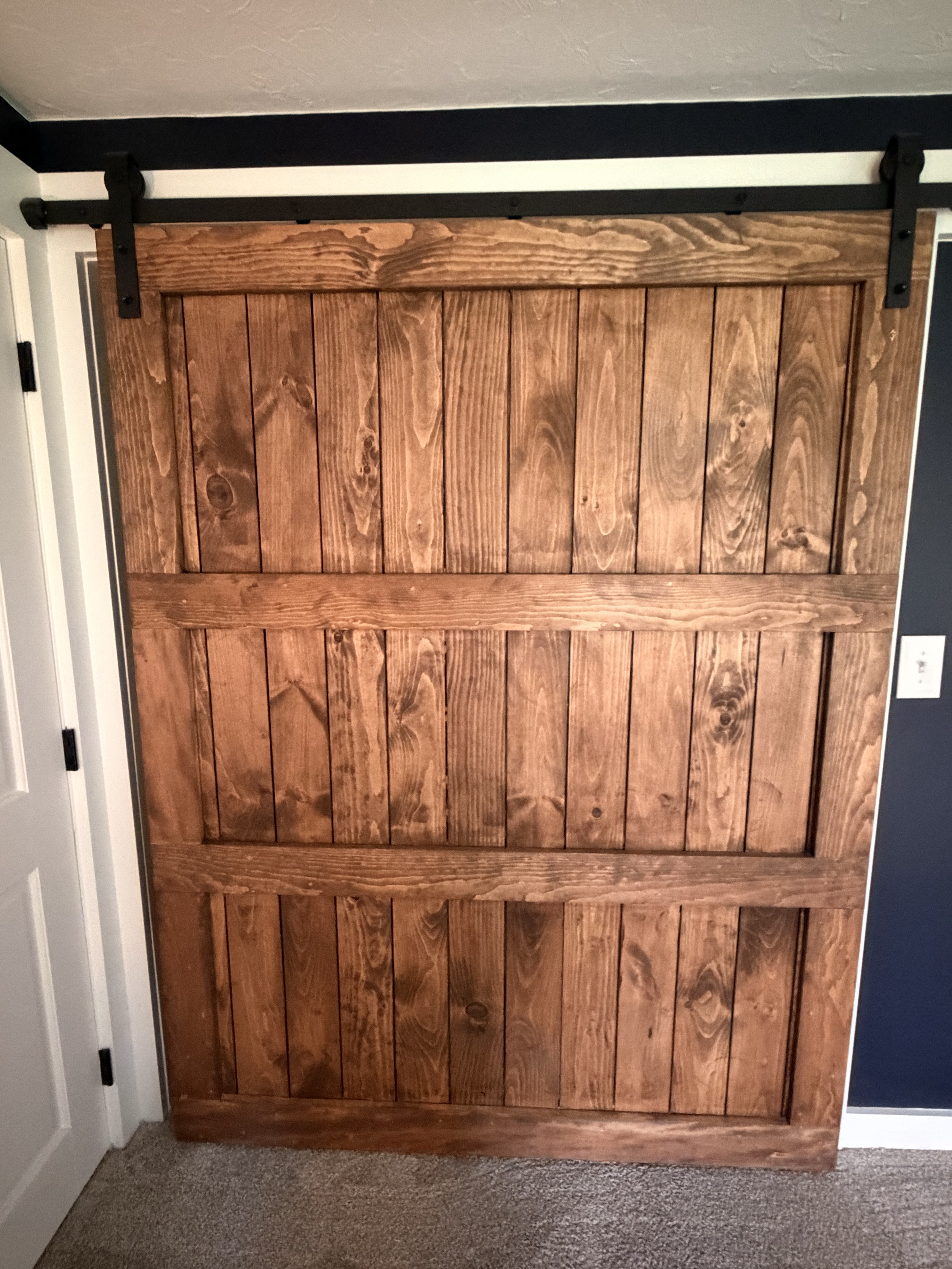 A wooden sliding barn door with black hardware on a wall in a room with a white door and gray walls.