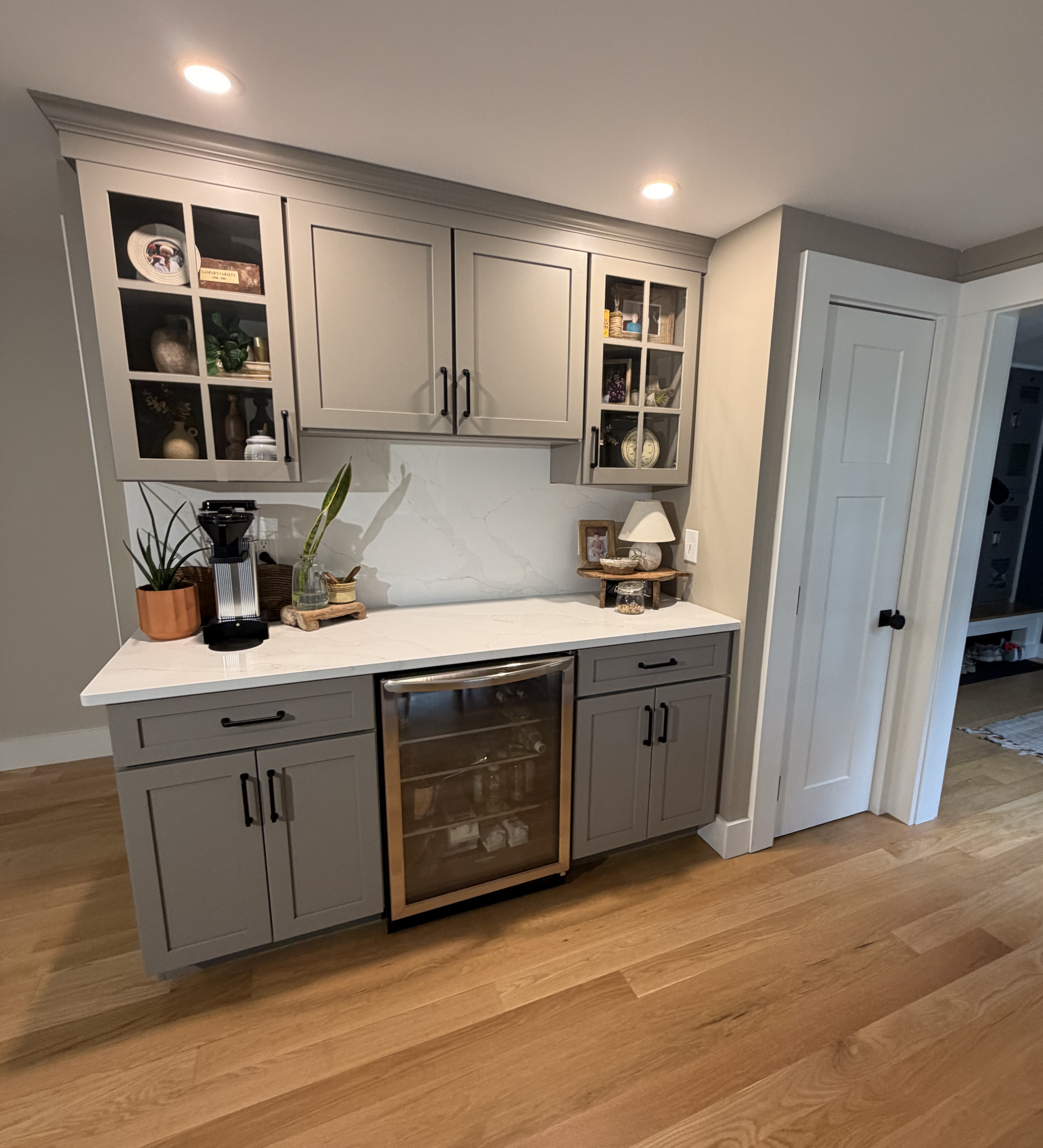 Gray kitchen cabinets with glass doors displaying decorative items, a white marble countertop, and a rustic wooden table below with plants and small decor items in front of the cabinets.