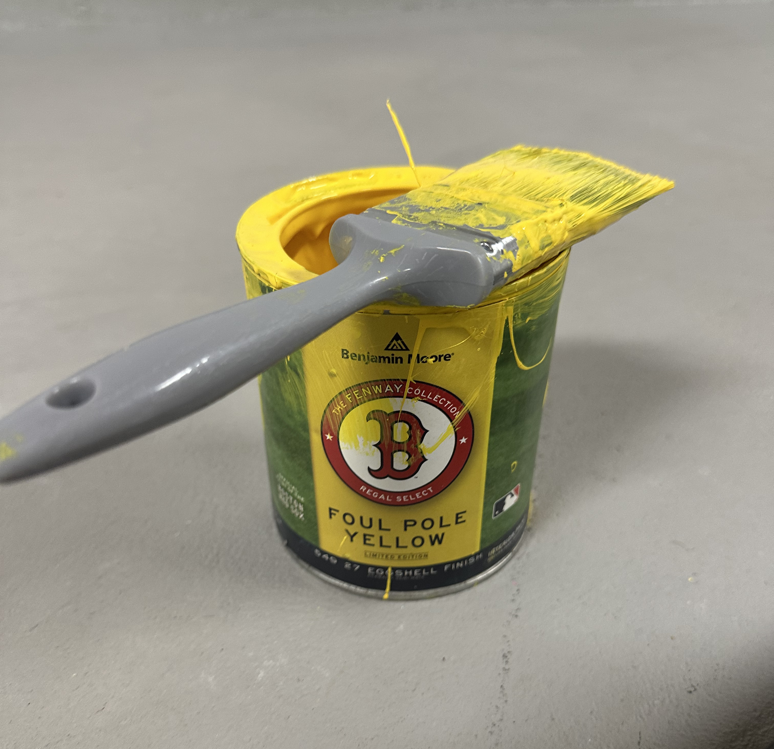 A partially opened tube of Benjamin Moore yellow paint with yellow paint smeared on the cap and the brush resting on top, set against a light gray background.