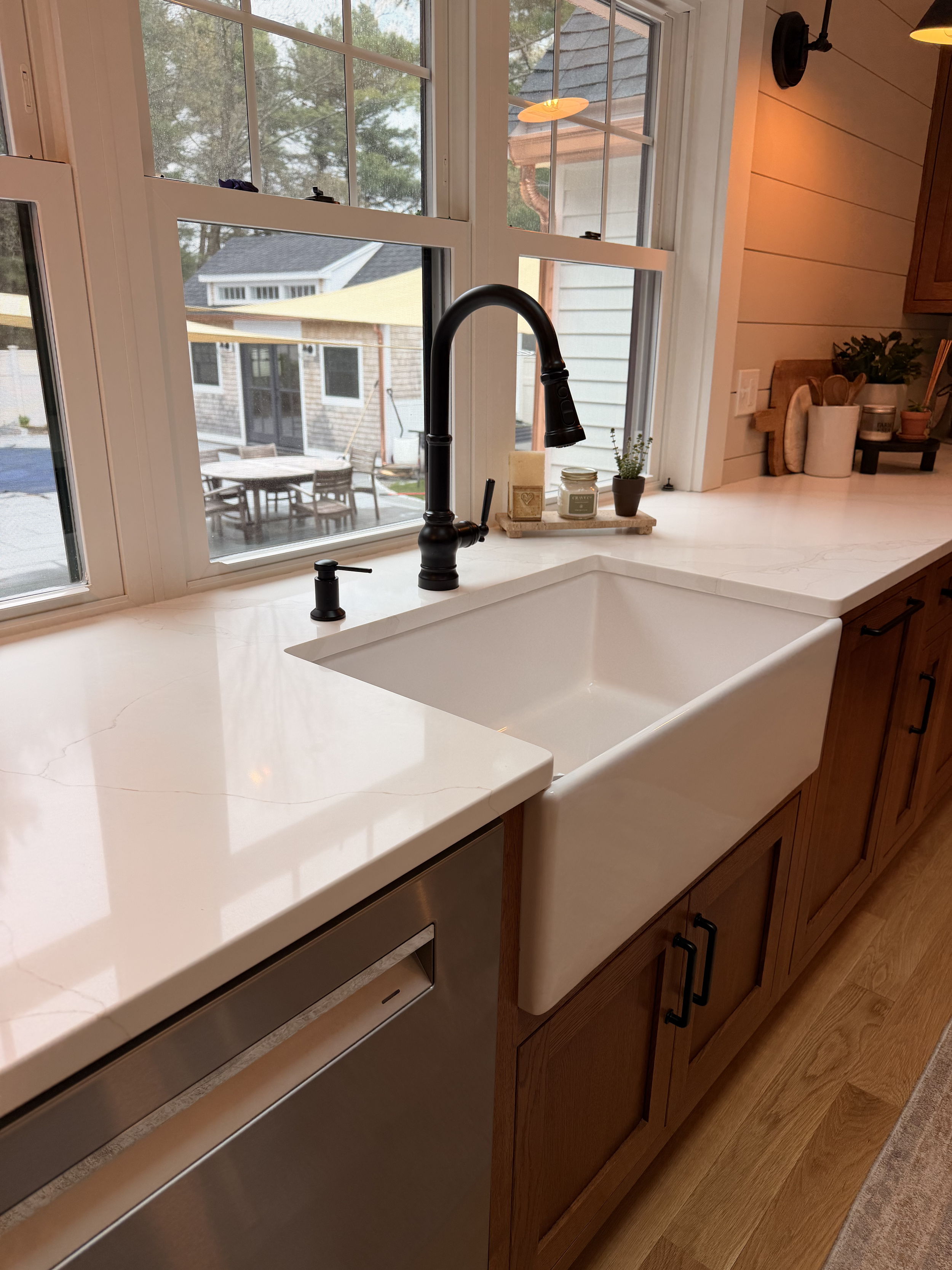Kitchen sink with black faucet, window above with outdoor patio furniture visible, copper-colored wood cabinets, beige countertop, small potted plant, candles on tray, wall-mounted speaker, and warm lighting.
