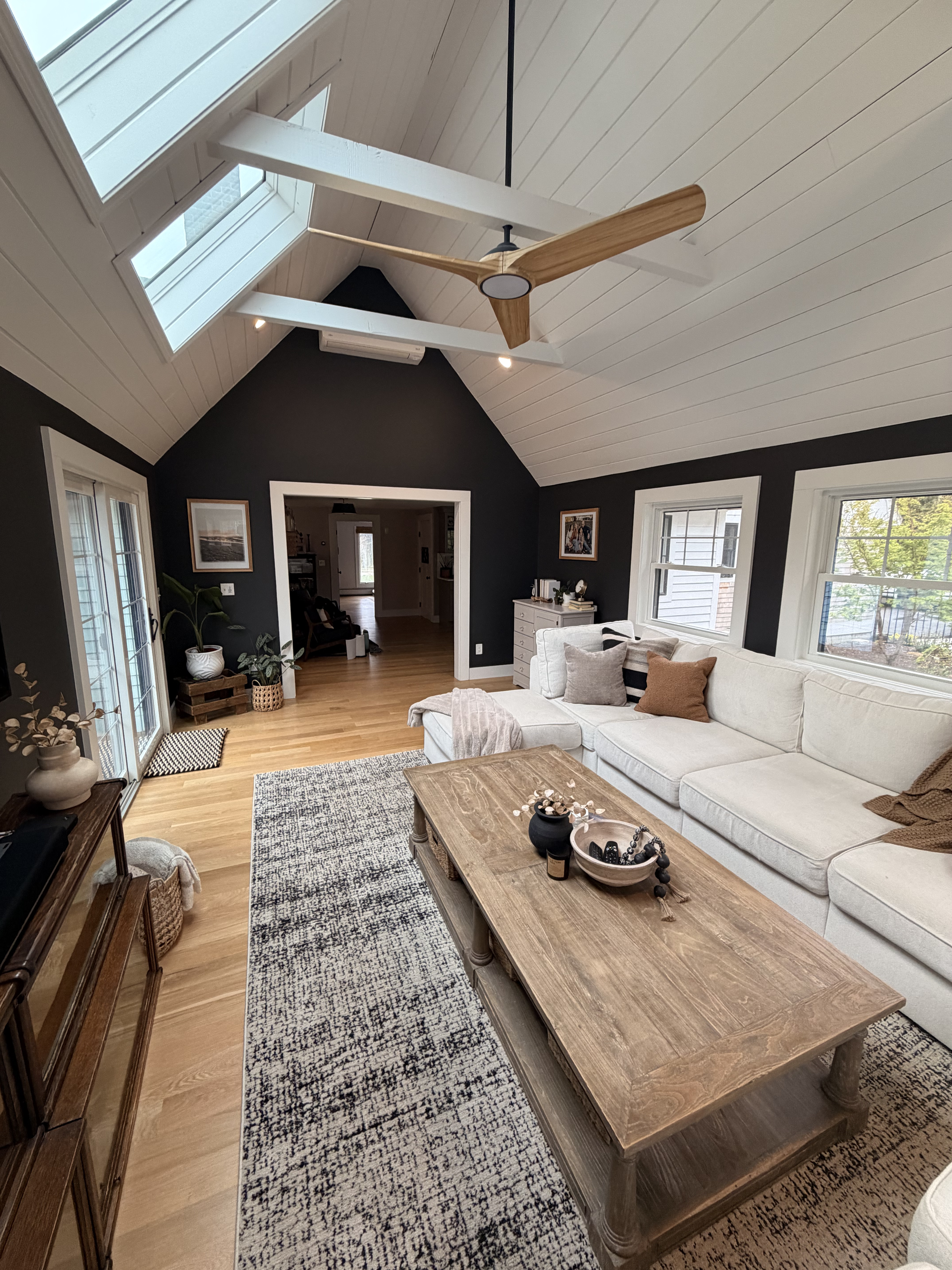 Living room with white sectional sofa, wooden coffee table, area rug, black and white walls, large windows, ceiling fan, framed art, potted plants, and adjacent dining area.