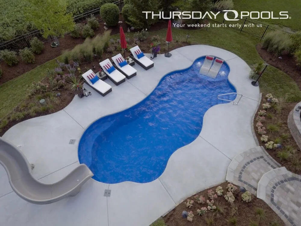 An aerial view of a backyard swimming pool with a slide, lounge chairs with striped towels, and some red umbrellas, surrounded by plants and a stone pathway.