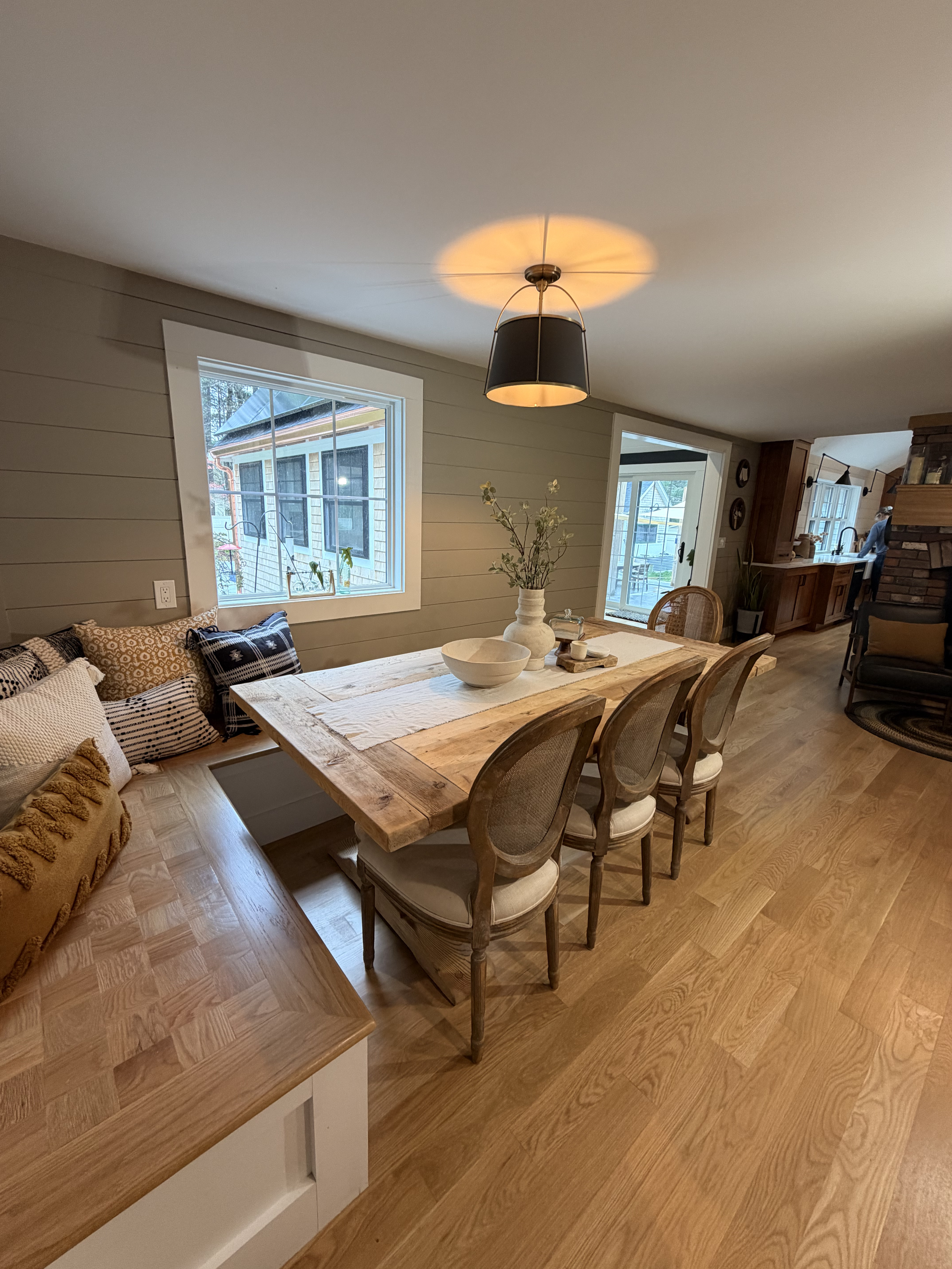 A cozy dining and living area with wooden flooring, a large wooden dining table, rattan chairs, a corner with a couch decorated with patterned cushions, a window letting in natural light, and a view of a porch and other houses outside.