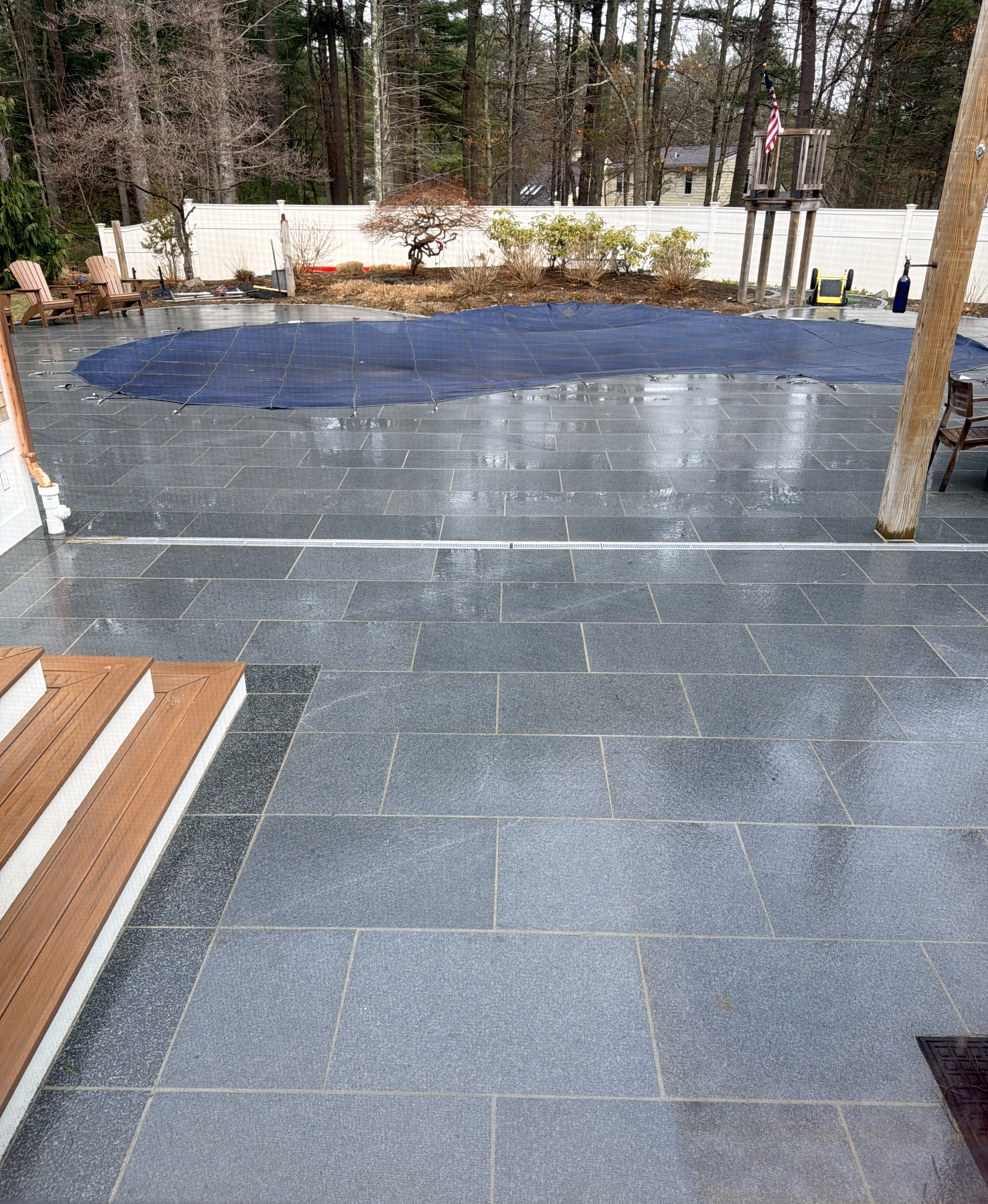 Backyard patio with black tiles, a blue pool cover, wooden outdoor chairs, a small deck with an American flag, a firepit, and several plants and trees in the background.