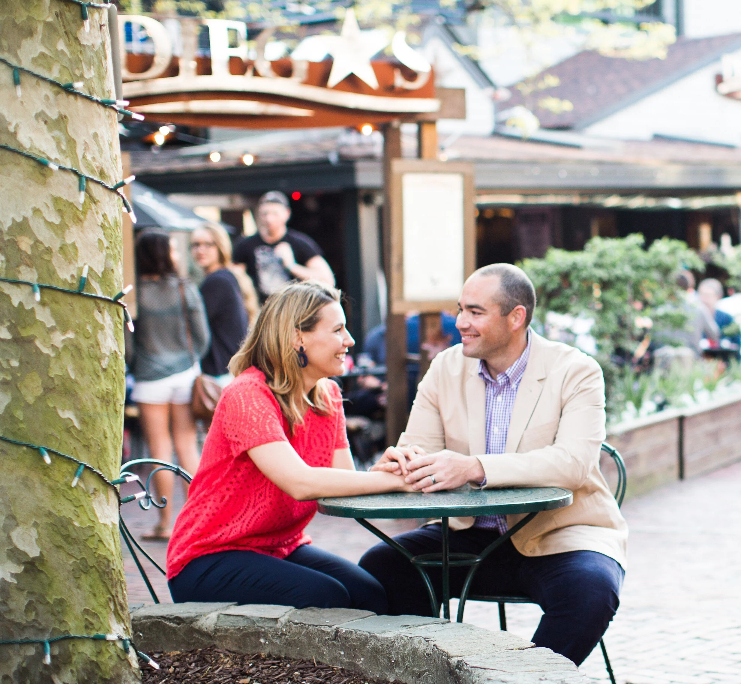 People sitting at a table outdoors, smiling and engaging in conversation, with a decorative tree in the foreground and a building in the background.