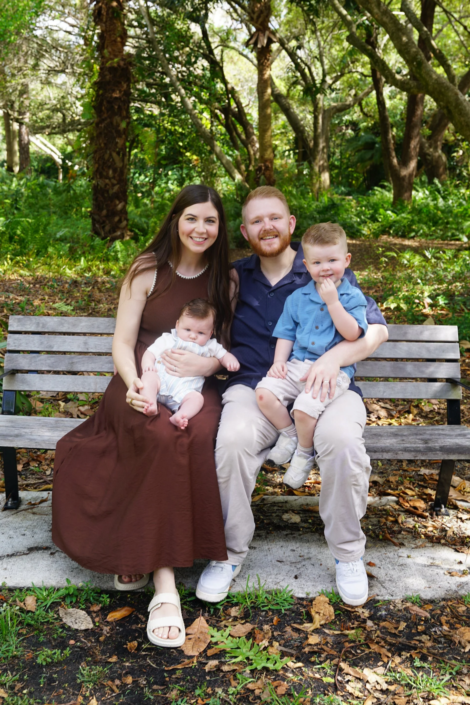 A family of four sitting on a park bench surrounded by trees and fallen leaves. The mother is holding a baby girl, the father is holding a young boy, and all are smiling at the camera.