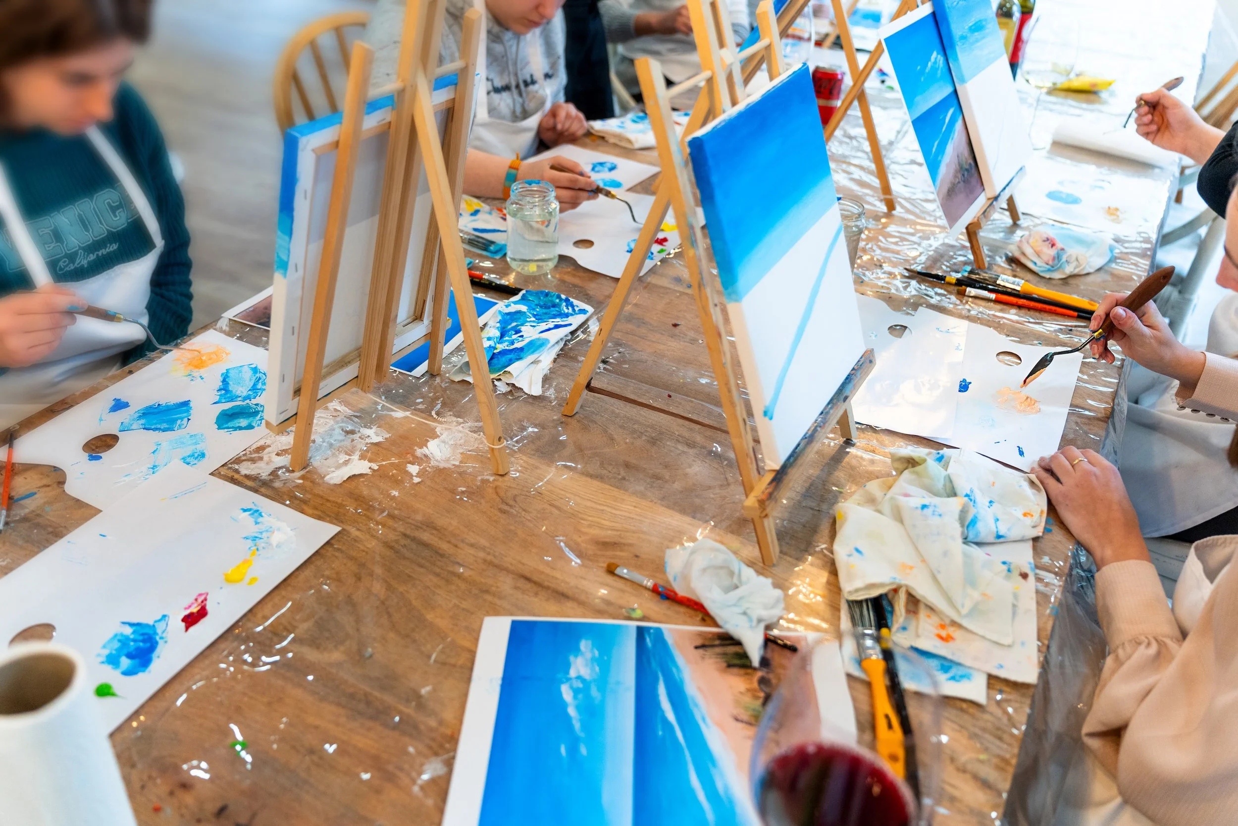 table with people sitting down at it with canvases, paint, and artbrushes