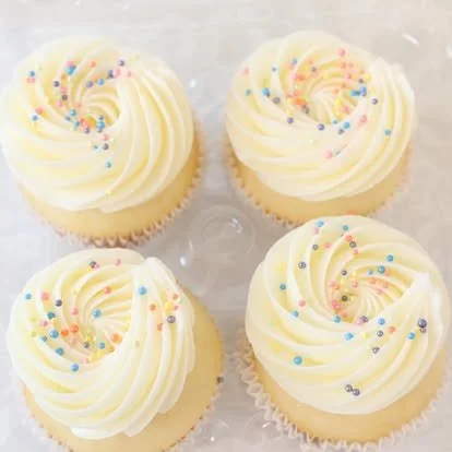 Four vanilla cupcakes with swirled white frosting topped with colorful round sprinkles.
