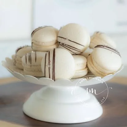 A white cake stand holding several macarons with white shells and dark chocolate drizzle.