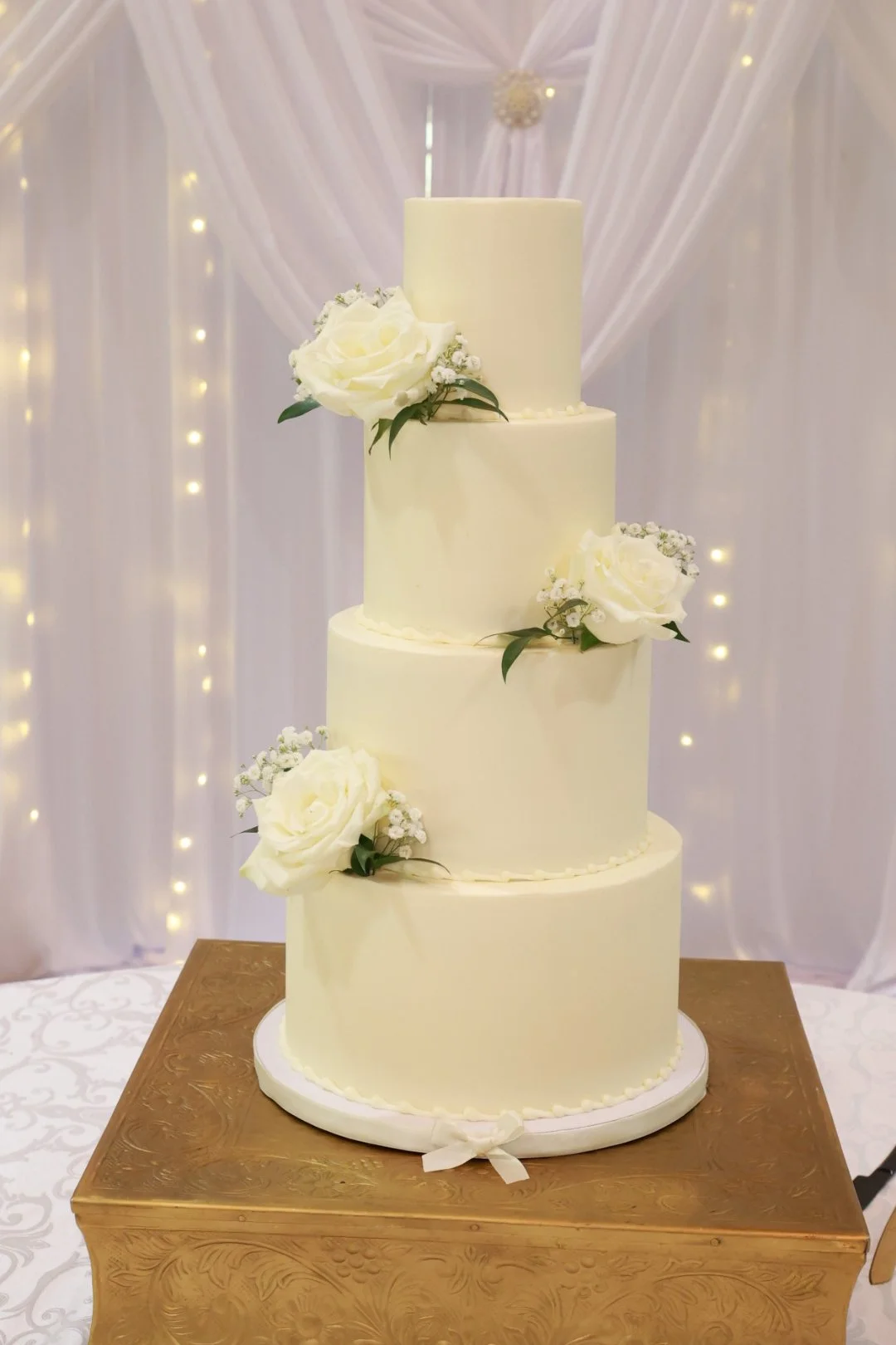 Four-tier white wedding cake decorated with white roses and greenery, placed on a wooden table.