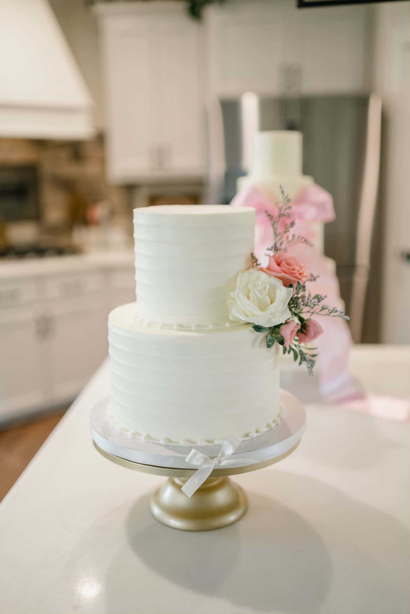 A three-tier white wedding cake decorated with pink and white flowers, placed on a decorative stand with a ribbon at the bottom.