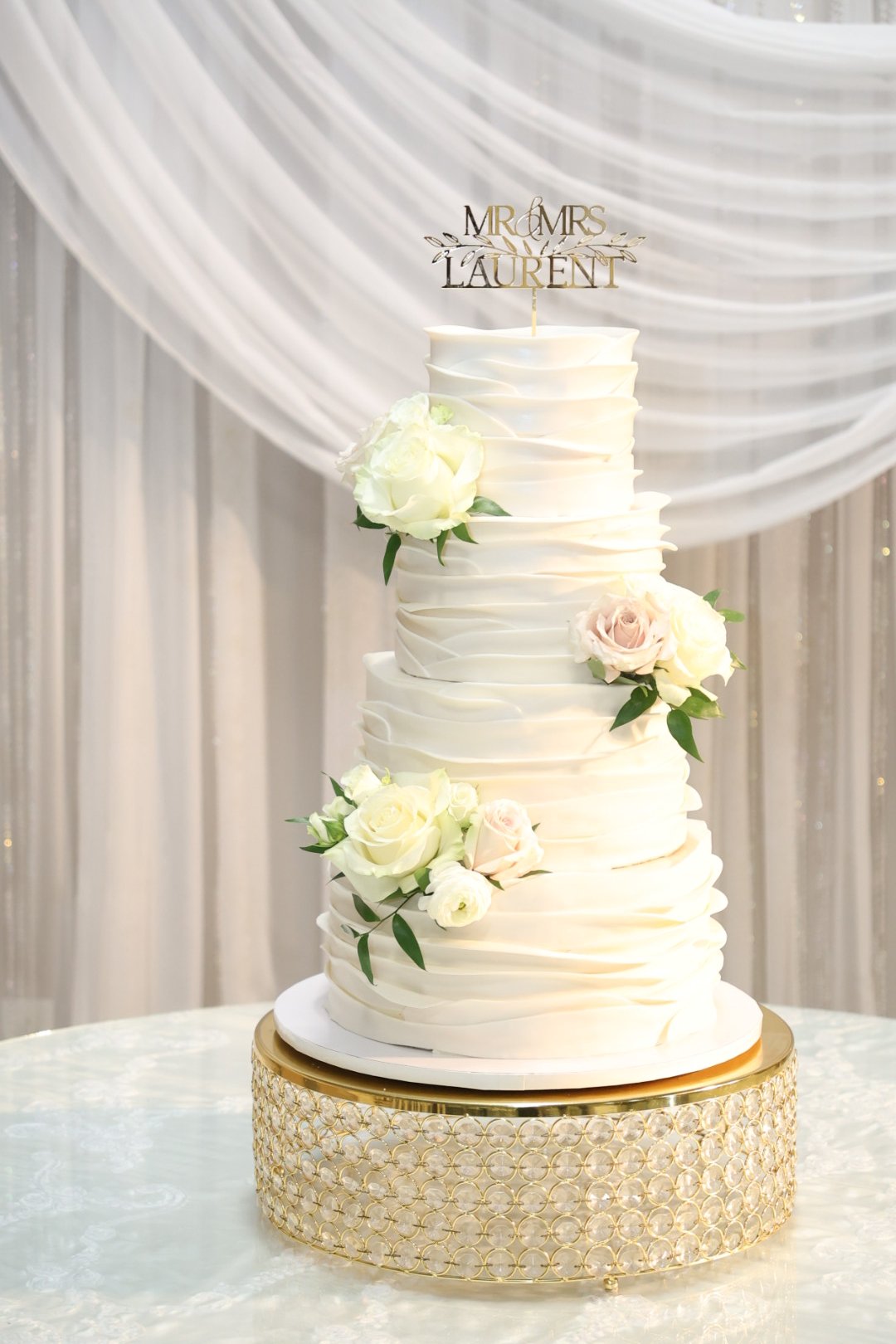 A tall, elegant wedding cake with a textured white frosting finish, decorated with white and blush roses and greenery, topped with a gold "MOMS LAURENT" cake topper, on a gold patterned cake stand.