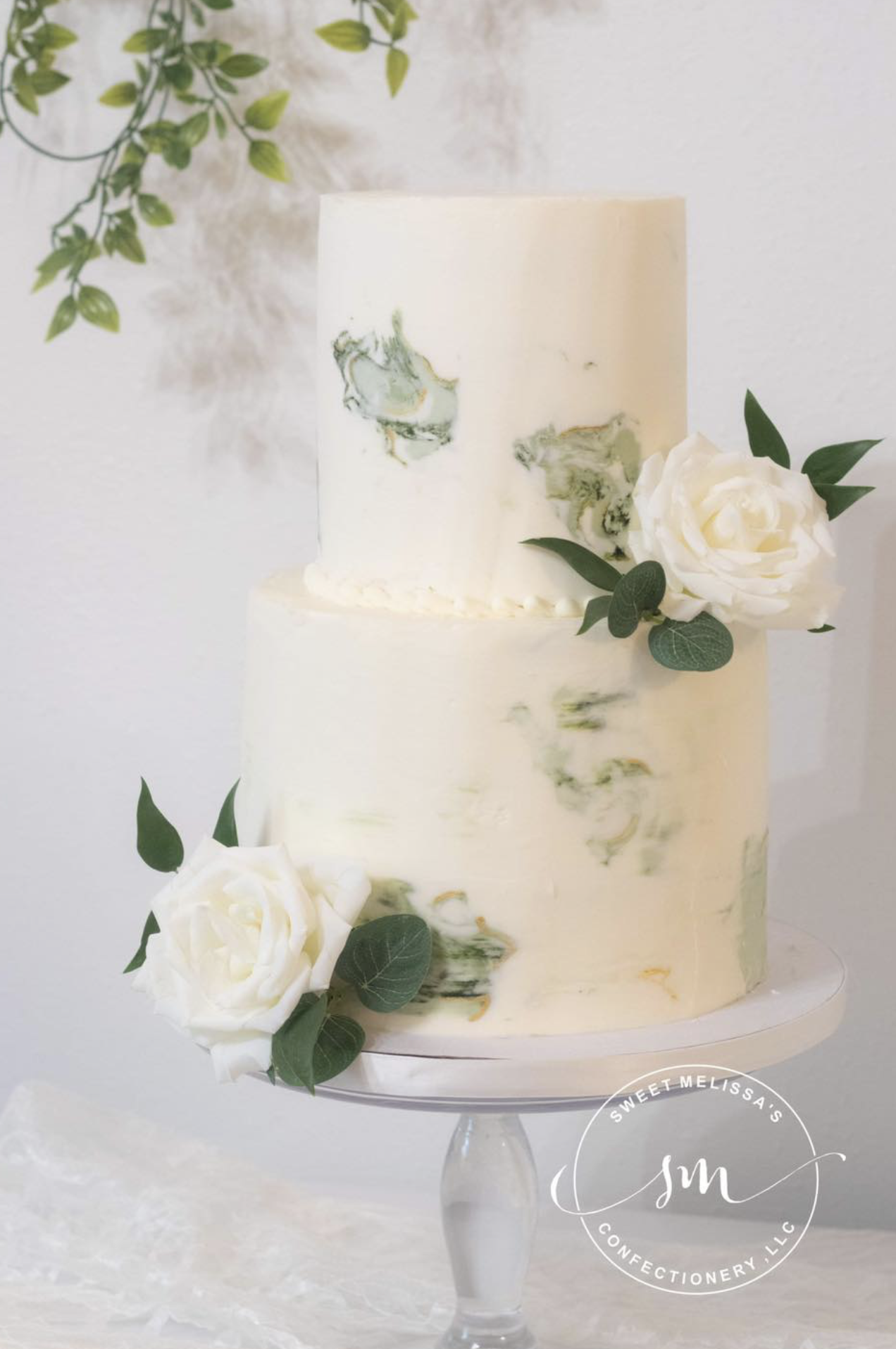 A two-tier white wedding cake decorated with white roses and green leaves, placed on a glass cake stand.
