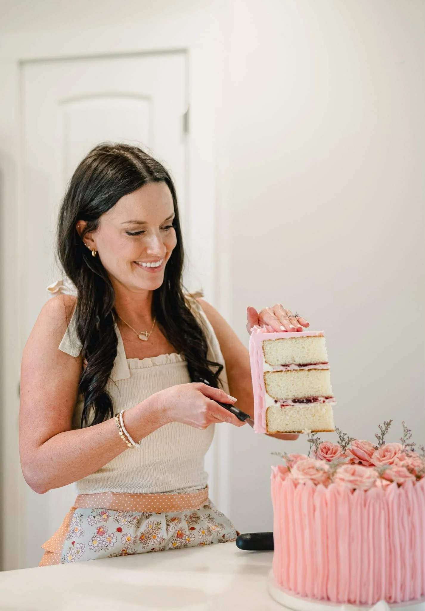 A woman with long dark hair, wearing a light-colored sleeveless top and an apron, is smiling while cutting a slice from a three-layer pink frosted cake. The cake has flower decorations on top and is placed on a white table, with a plain white background.