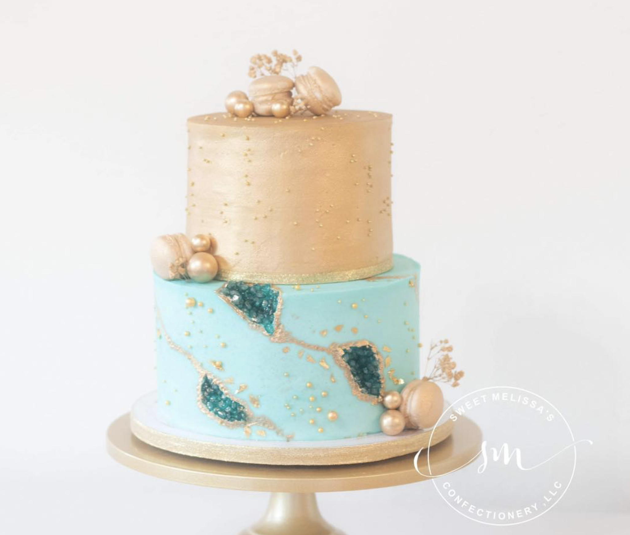 A two-tiered cake with a beige top layer decorated with gold and pearl accents, including gold beads and pearls, and a light blue bottom layer with gold veining and dark blue geode decoration. The cake is on a white cake stand against a plain white b