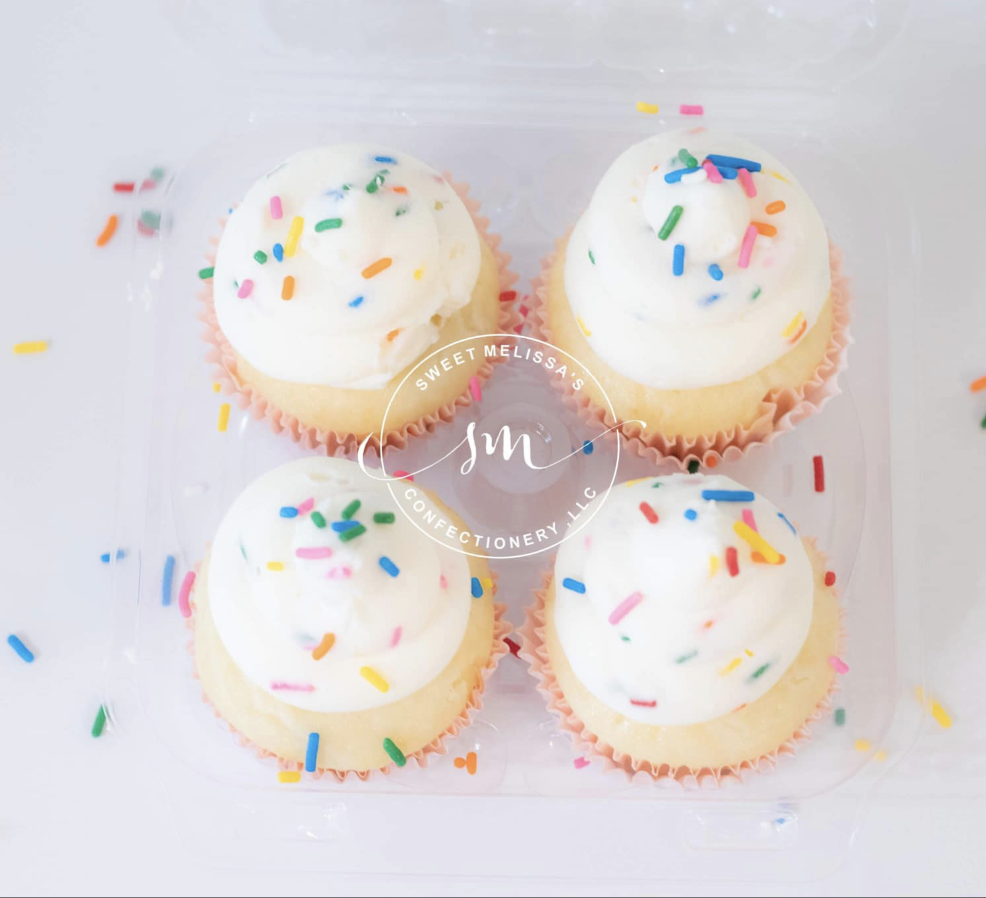 Two cupcakes with white icing and colorful sprinkles in pink wrappers on a white surface. 