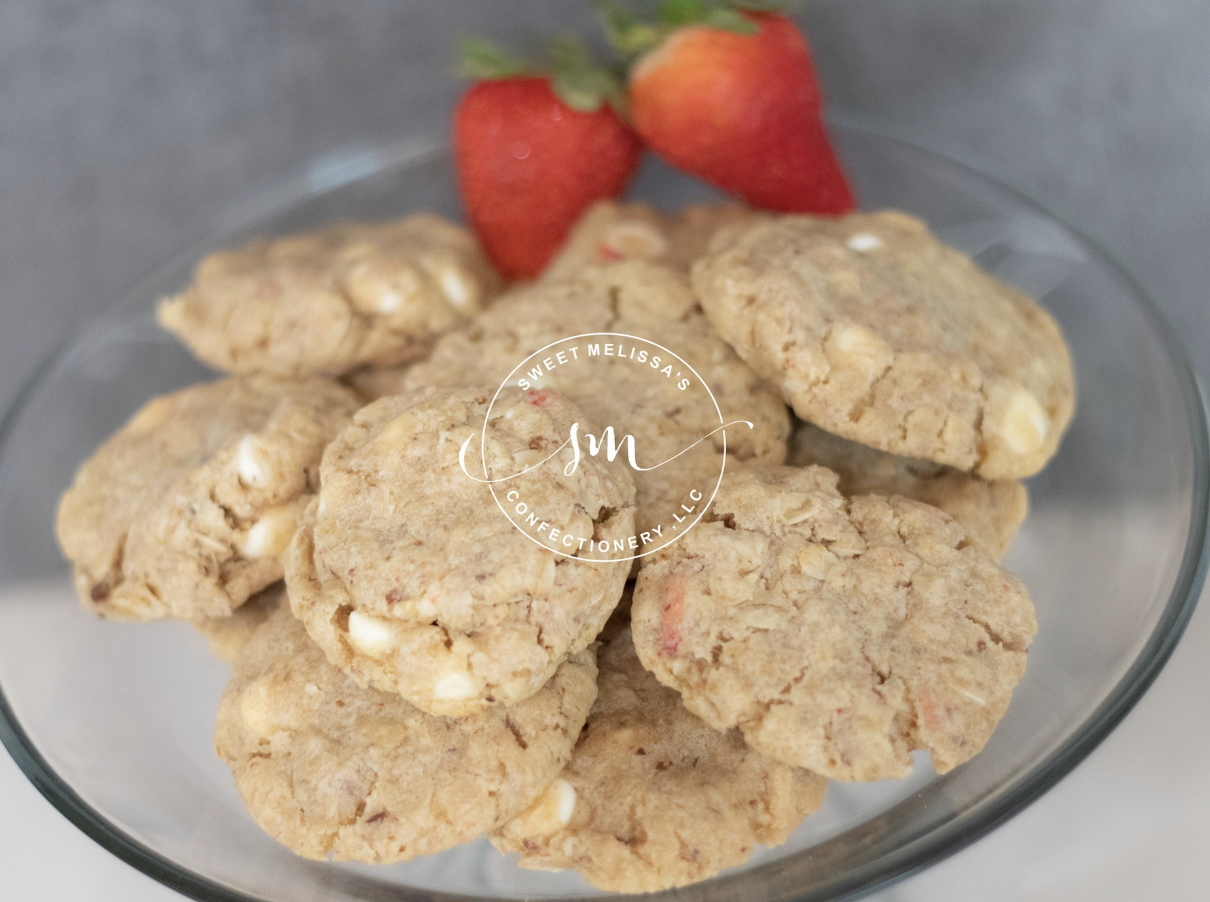 Close-up of a glass bowl containing oatmeal cookies with white chocolate chips and pieces of freeze-dried strawberries, topped with two fresh strawberries.