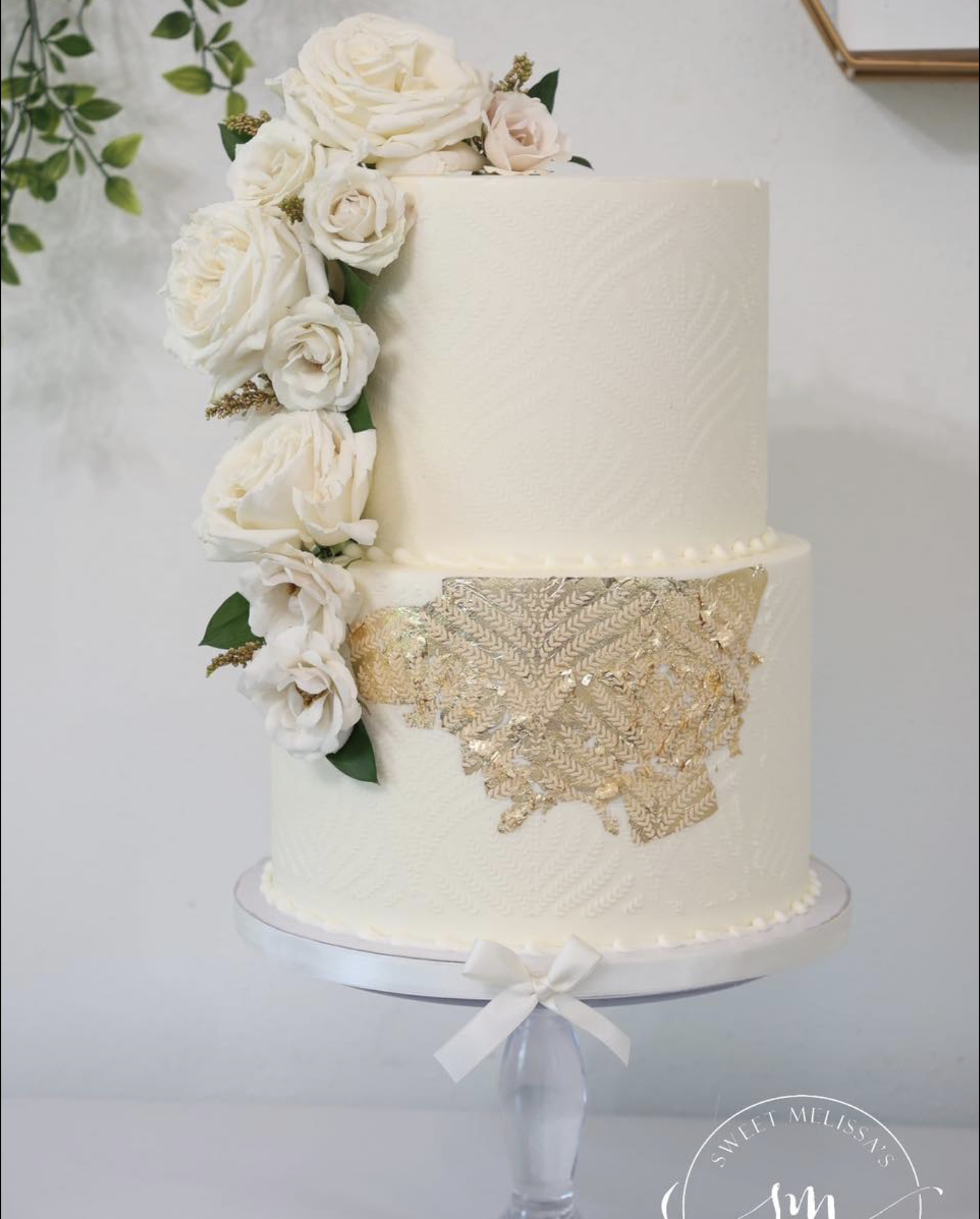 A two-tiered wedding cake decorated with white roses and greenery, and gold lace accents, on a stand with a ribbon around its base.