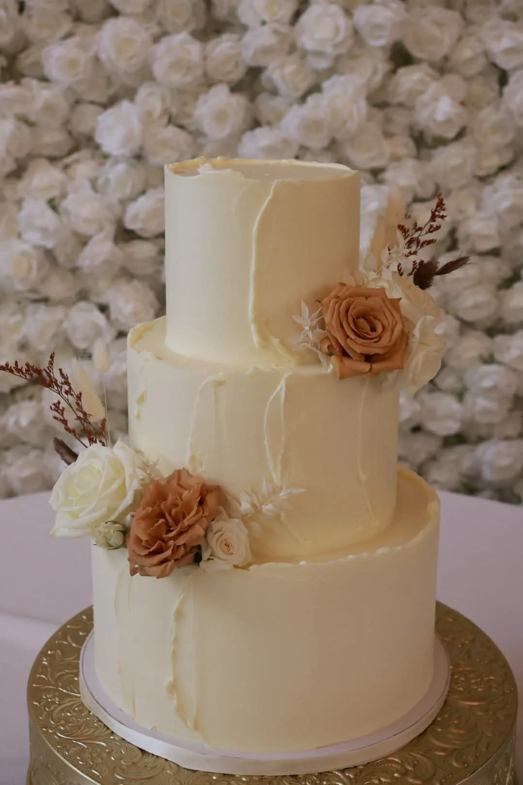 Three-tiered wedding cake decorated with cream flowers and beige roses, with a floral backdrop.