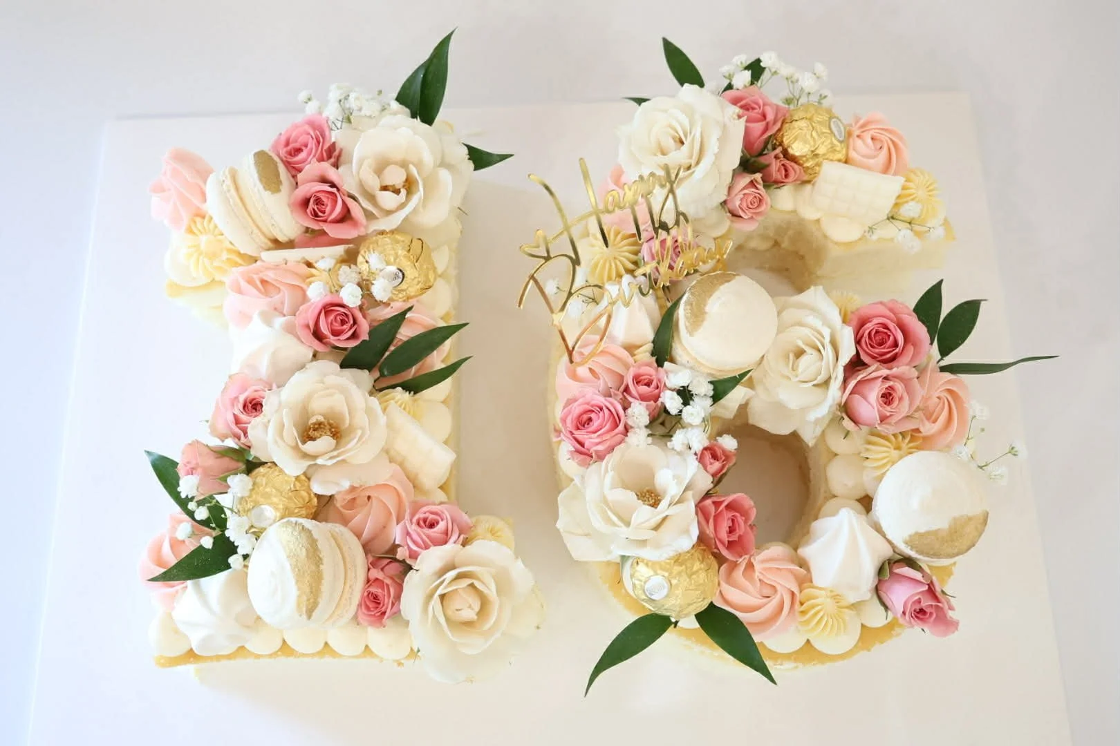 A white birthday cake shaped as the number 10, decorated with pink and white flowers, gold-wrapped chocolates, macarons, small white flowers, and green leaves, with a gold 'Happy Birthday' topper.