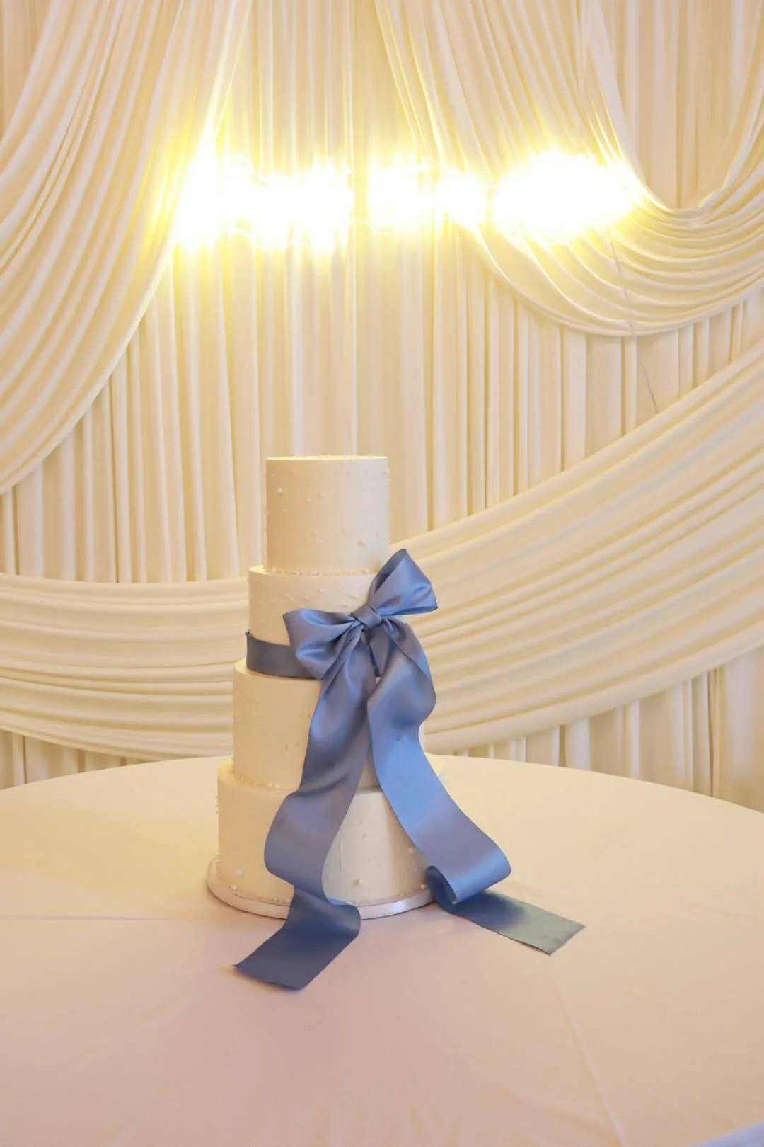 A three-tiered white wedding cake with a blue ribbon and bow, set on a round table in front of cream-colored curtains with warm lighting.