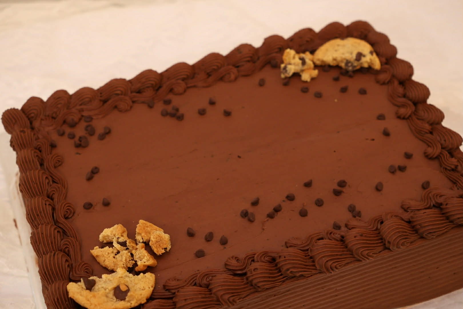 A rectangular chocolate cake with piped chocolate frosting border, small chocolate chips on top, and crumbled cookie pieces on the corners.