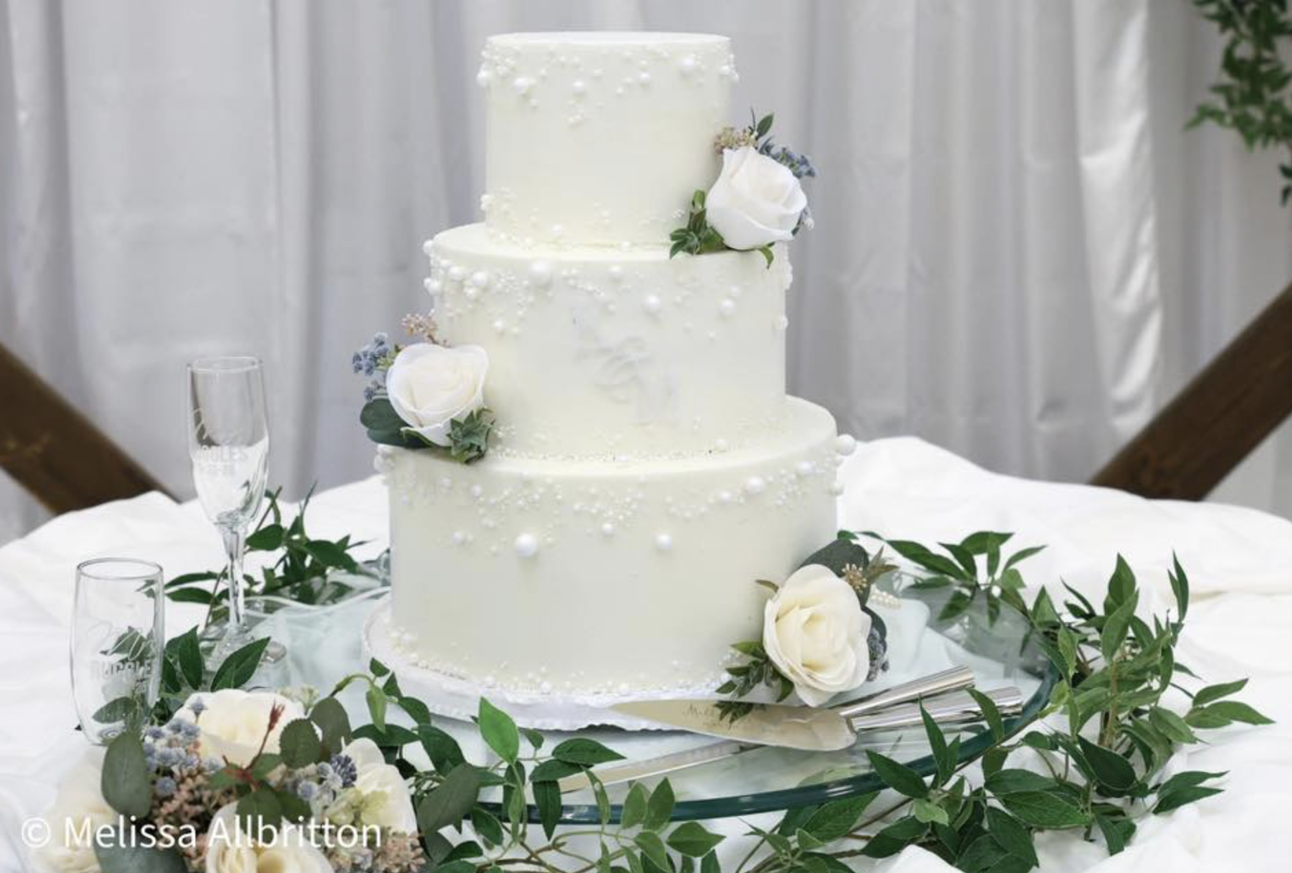 Three-tier white wedding cake decorated with white flowers and small white pearls on top of a decorated table with greenery and white flowers around it.