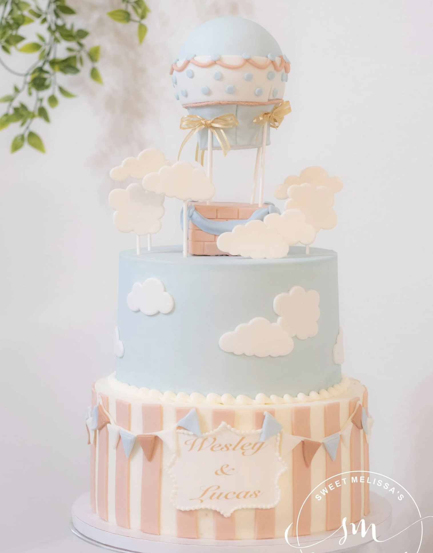 A pastel pink and blue tiered cake with clouds and bunting decorations, featuring a hot air balloon topper with polka dot and ribbon designs.